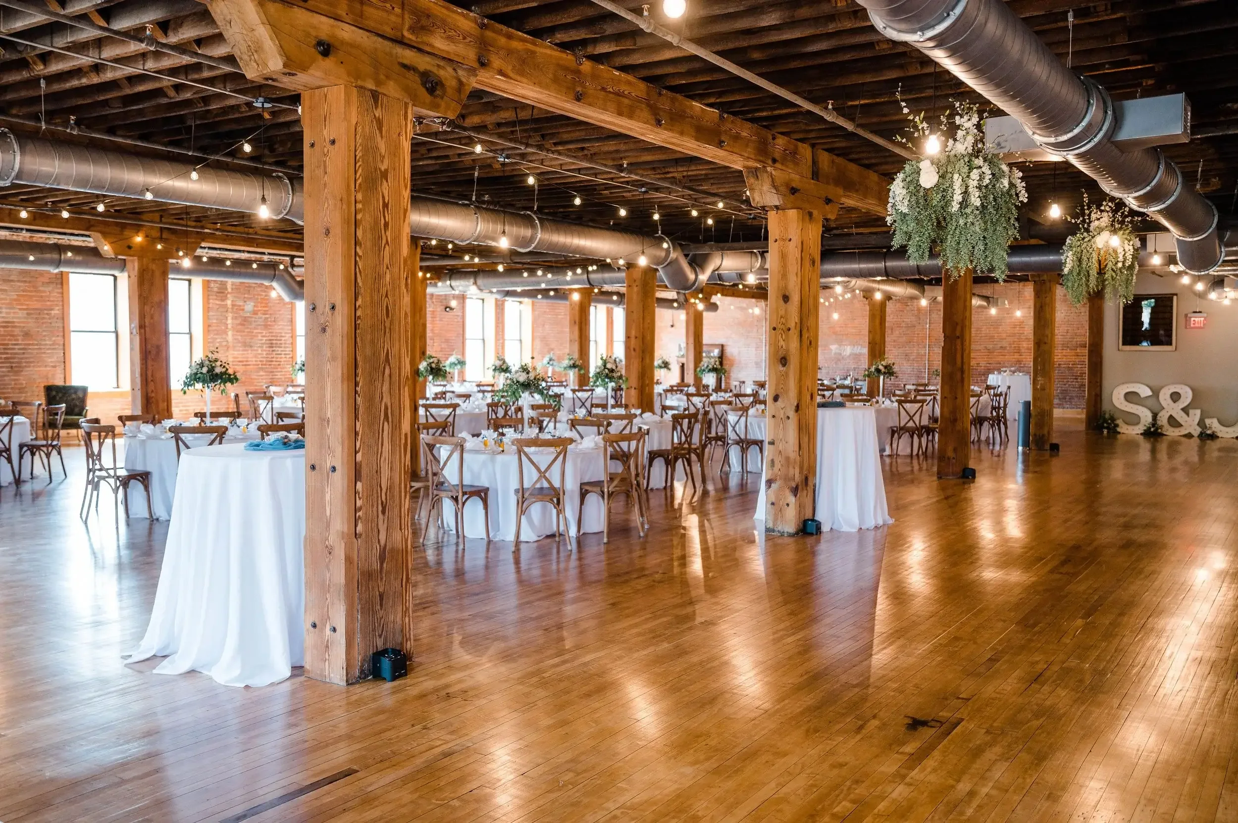 An indoor event space with wooden floors, exposed brick walls, large support beams, potted plants, and tables with white tablecloths, chairs, and floral centerpieces, decorated with hanging string lights and greenery.