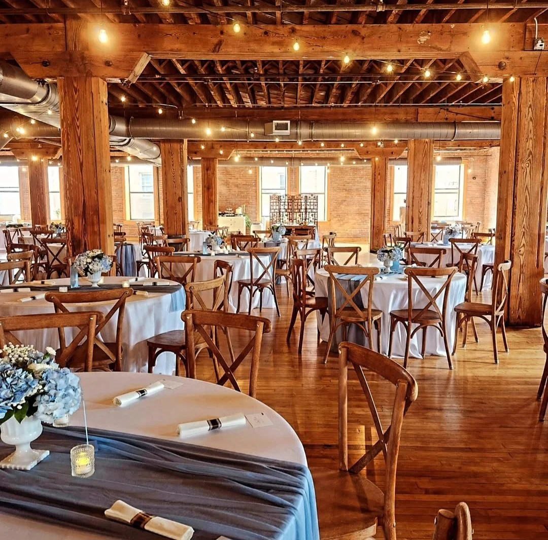 A spacious banquet hall decorated for a wedding or event, featuring round tables with white tablecloths, floral centerpieces, and wooden chairs. The hall has exposed brick walls, large windows, wooden beams, and string lights hanging from the ceiling