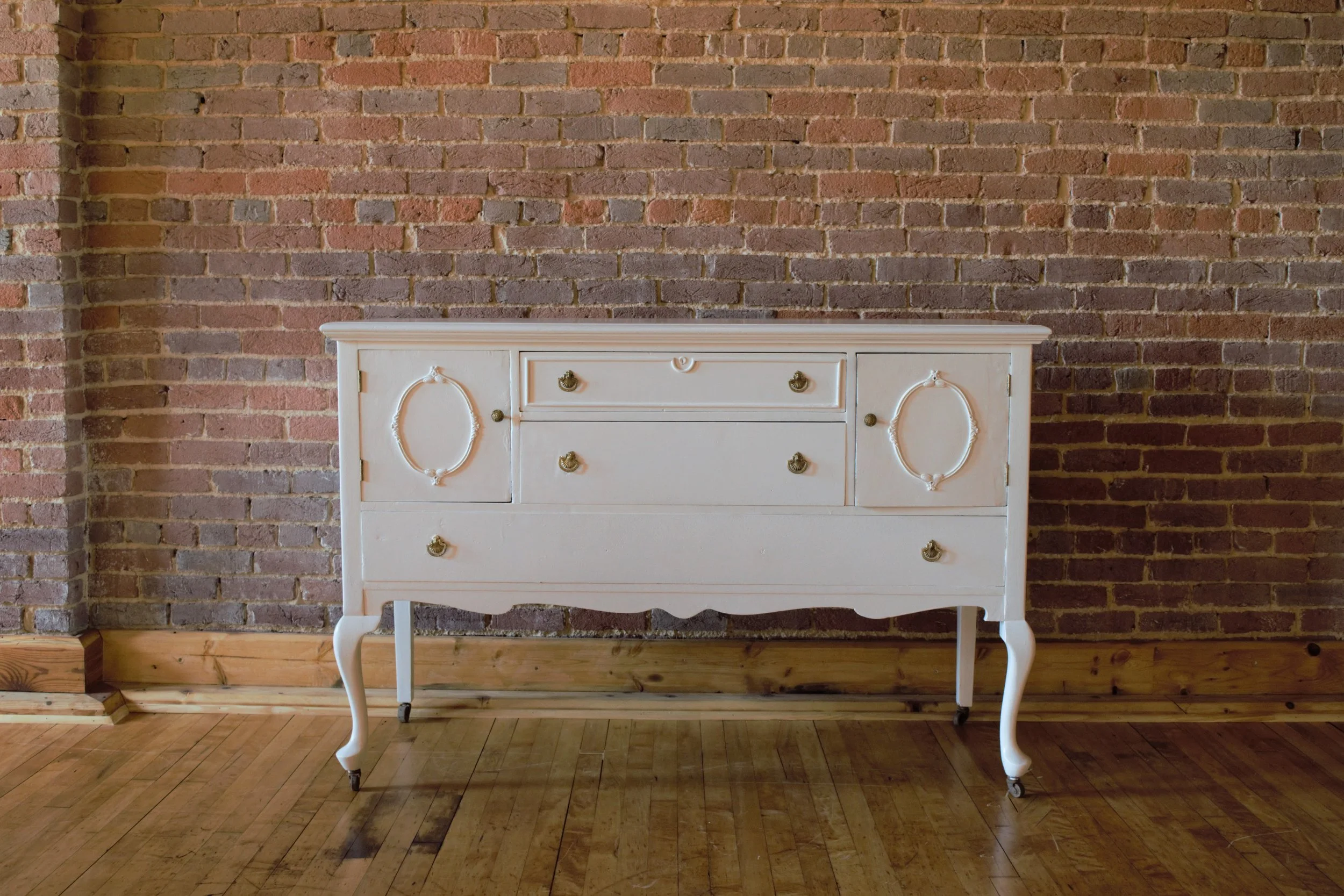A vintage white dresser with ornate details and brass handles, standing on carved legs with small wheels, placed against an exposed brick wall on a wooden floor.