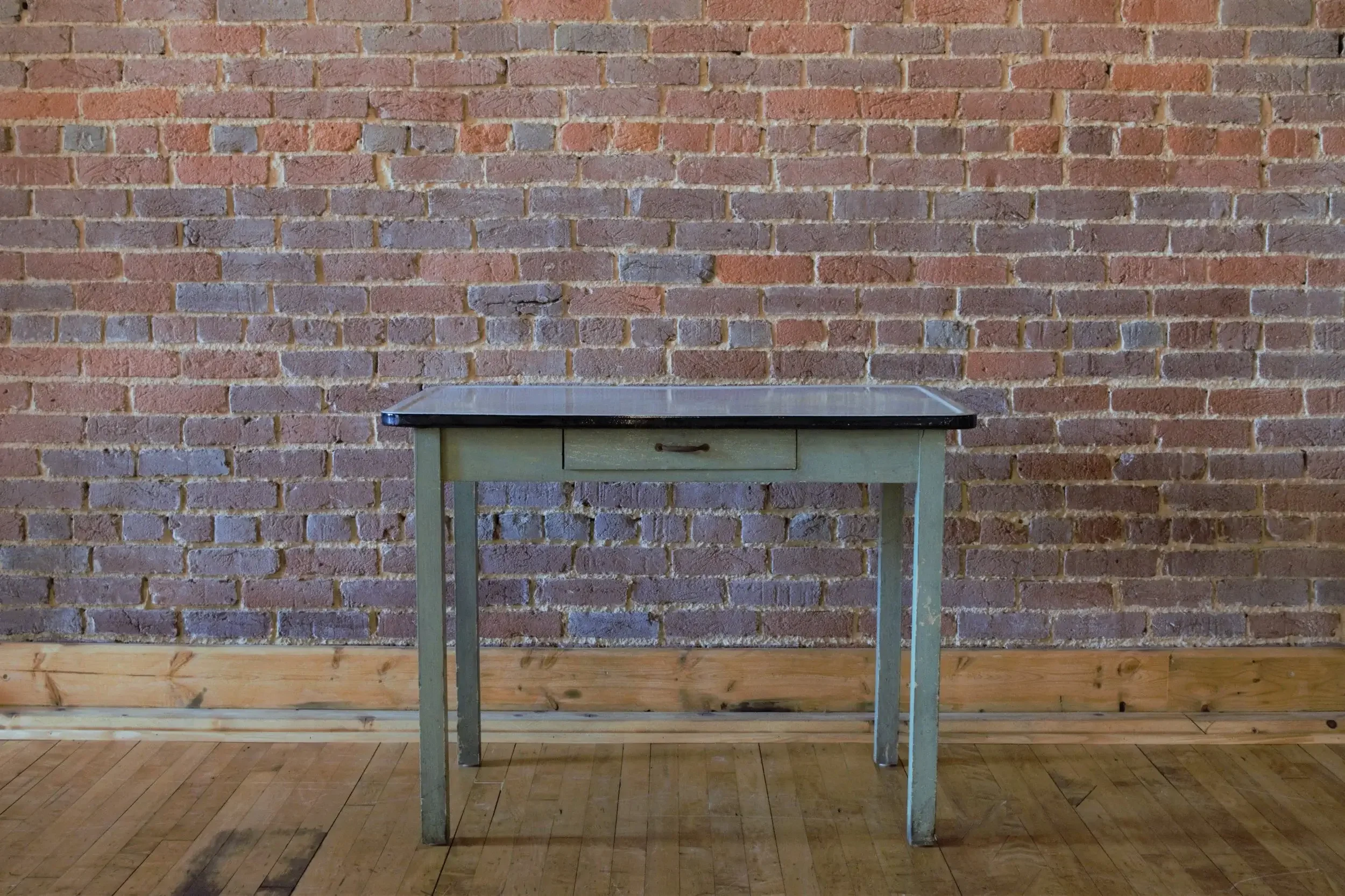 Small vintage green wooden table with a black top and a drawer, set against a brick wall with a wooden floor.