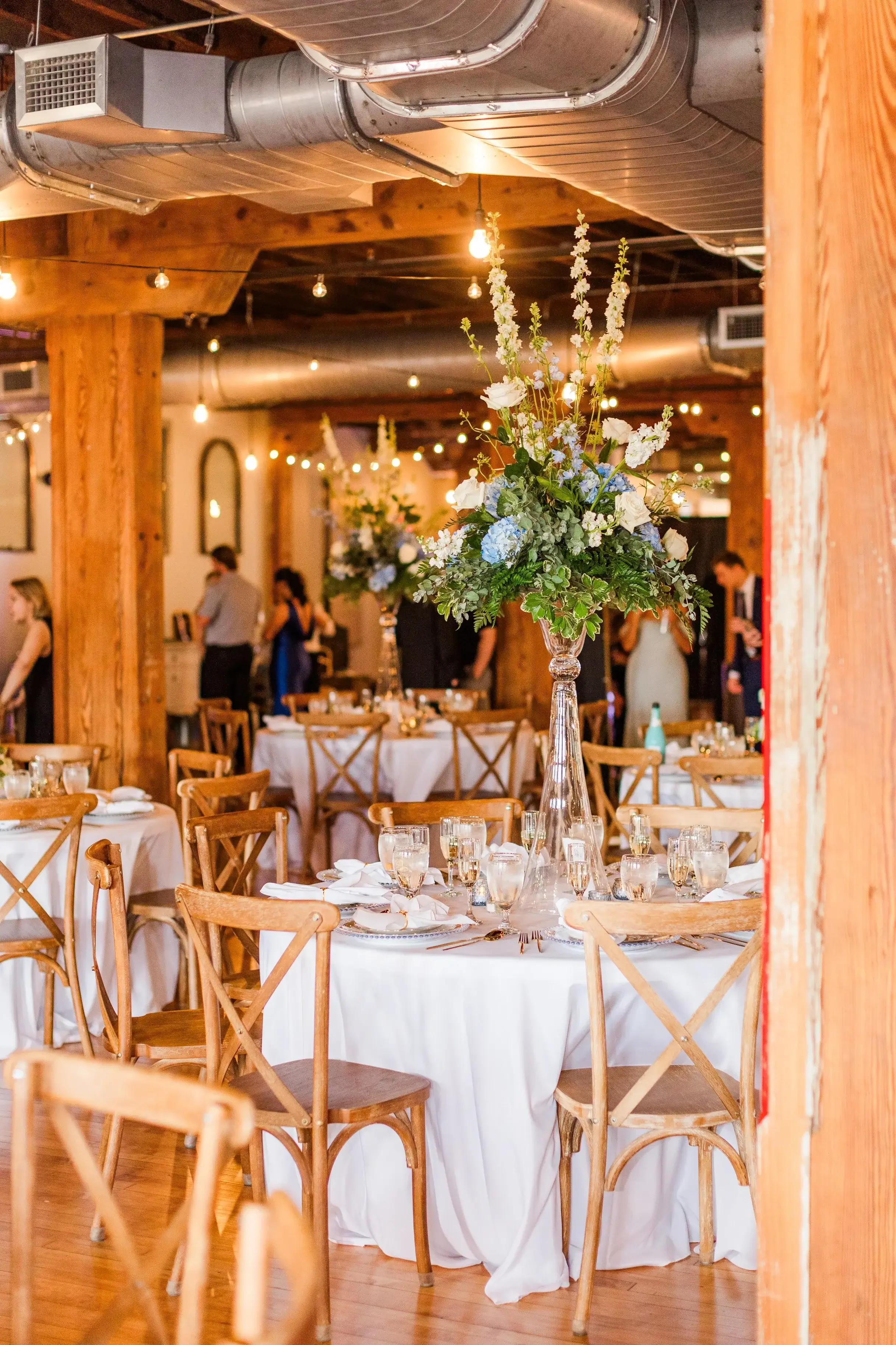 Elegant event setup with round tables decorated with white tablecloths and large floral centerpieces, wooden chairs, and glassware, inside a rustic venue with exposed ductwork and string lights.