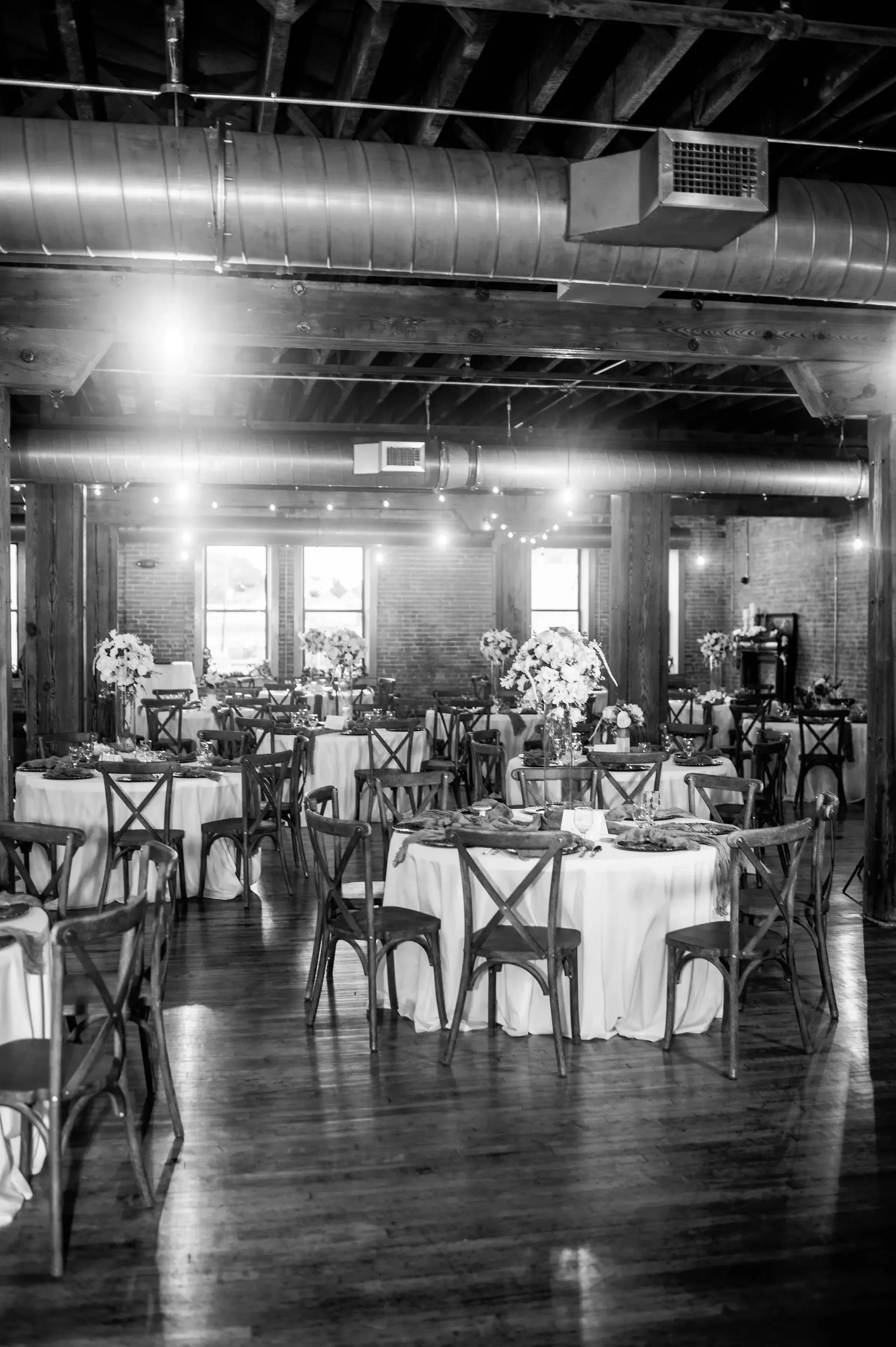 Black and white photo of a decorated event space with round tables draped in white cloth, floral centerpieces, and wooden chairs, in a rustic-style room with exposed brick walls, wooden beams, and large windows.