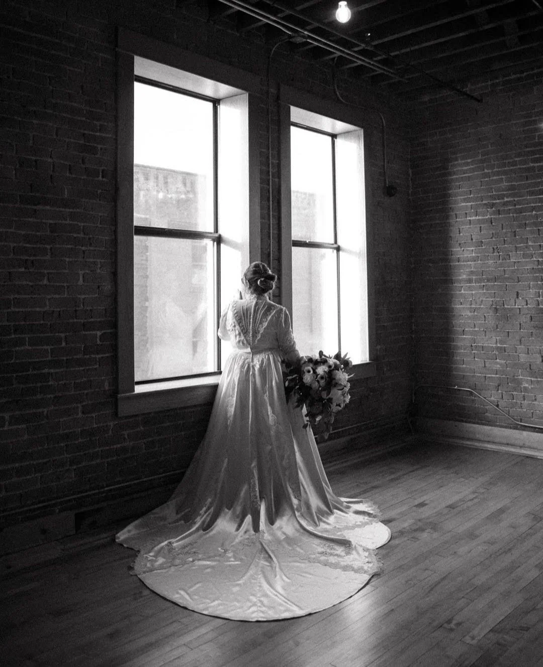 A woman in a wedding dress holding a bouquet, standing by a large window in a brick-walled room, with soft light illuminating the scene in black and white.