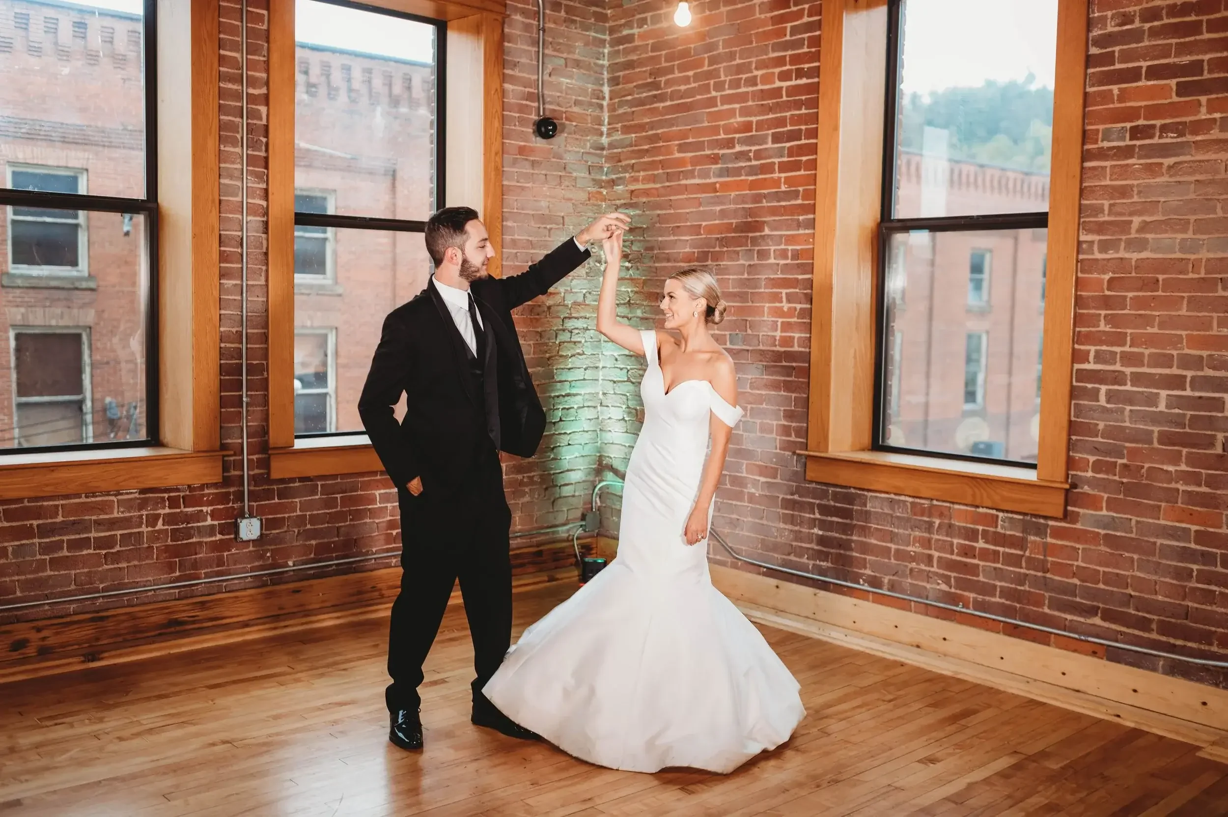 A bride and groom dancing in a spacious, industrial-style room with exposed brick walls and large windows