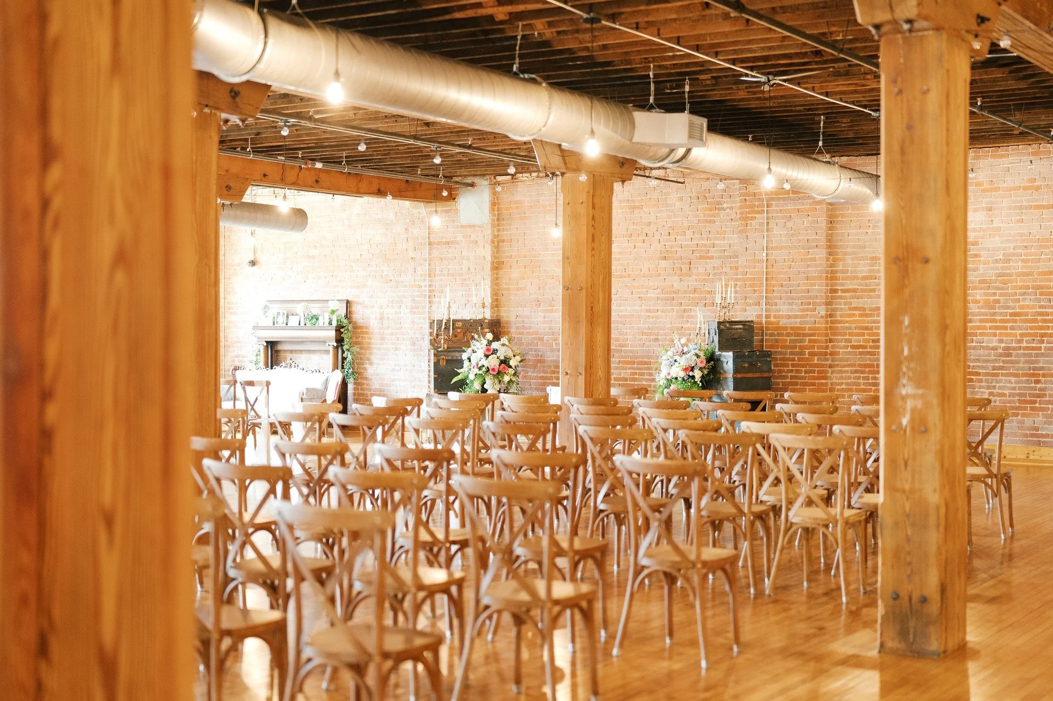 An event space with wooden chairs arranged facing a decorated area with flowers and black trunks, exposed brick walls, wooden support beams, and string lights.