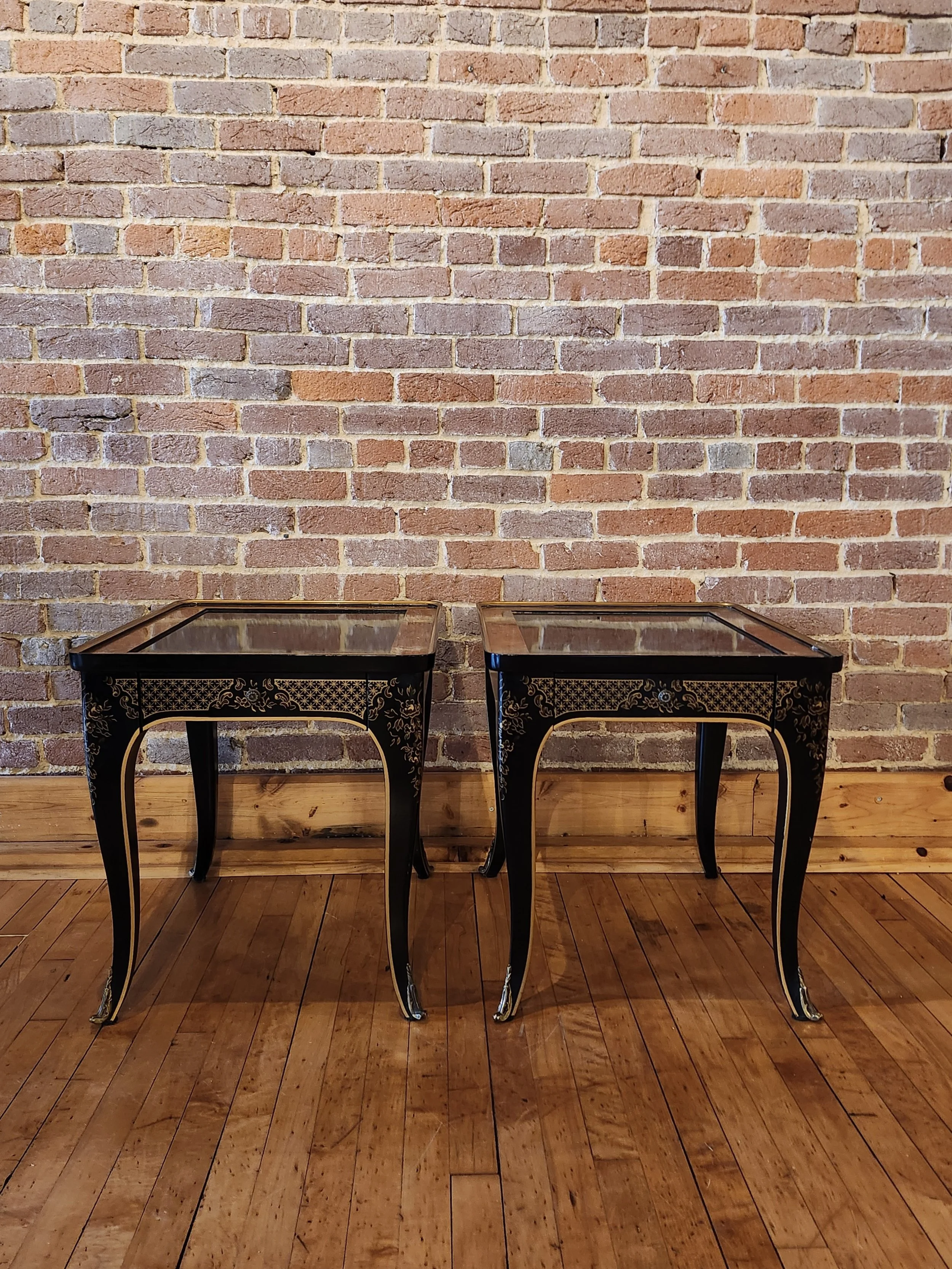 Two black decorative tables with curved legs and gold accents, placed side by side on a wooden floor against a brick wall background.