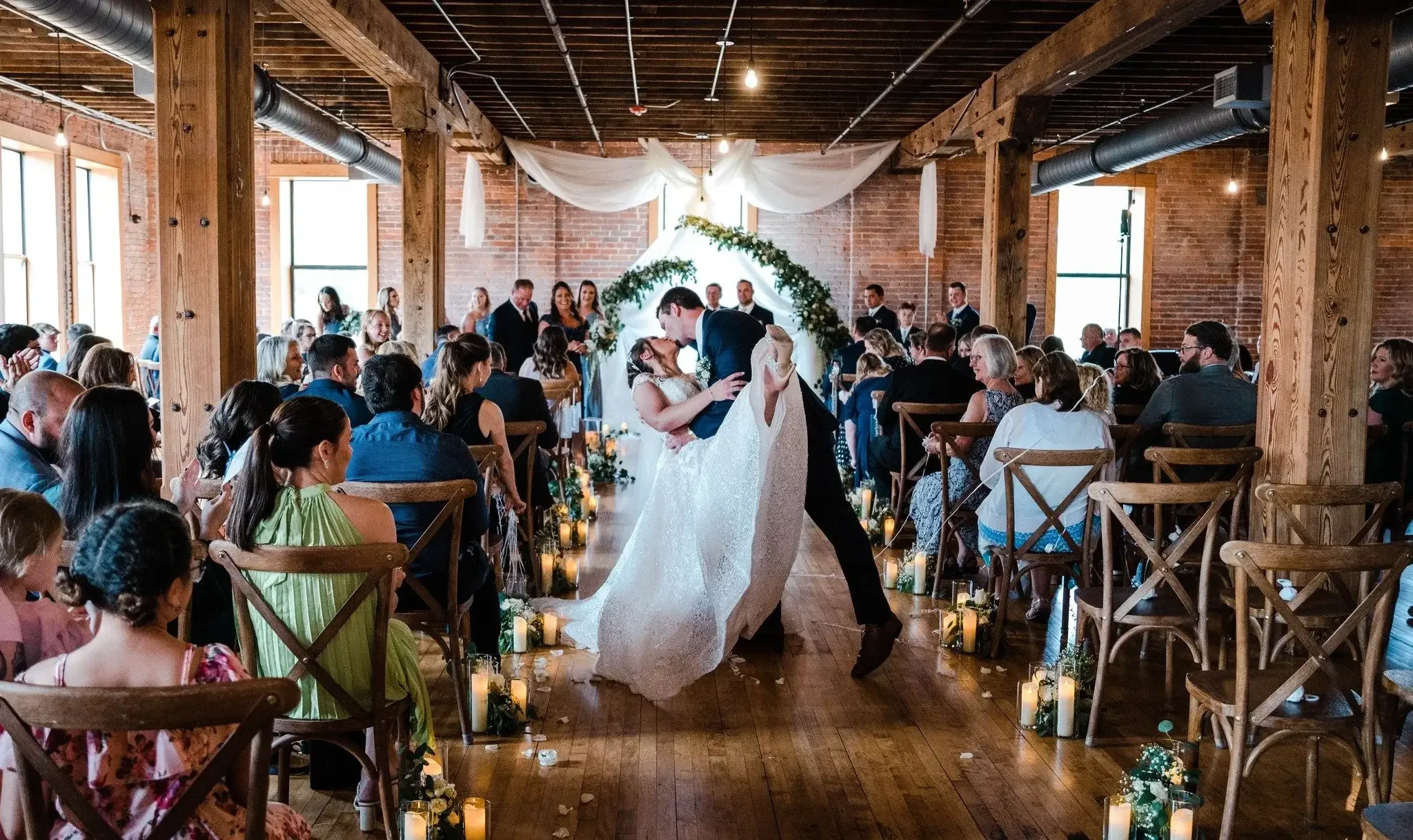 A couple dancing at their wedding reception in a rustic indoor venue with brick walls and wooden beams, surrounded by seated guests watching.