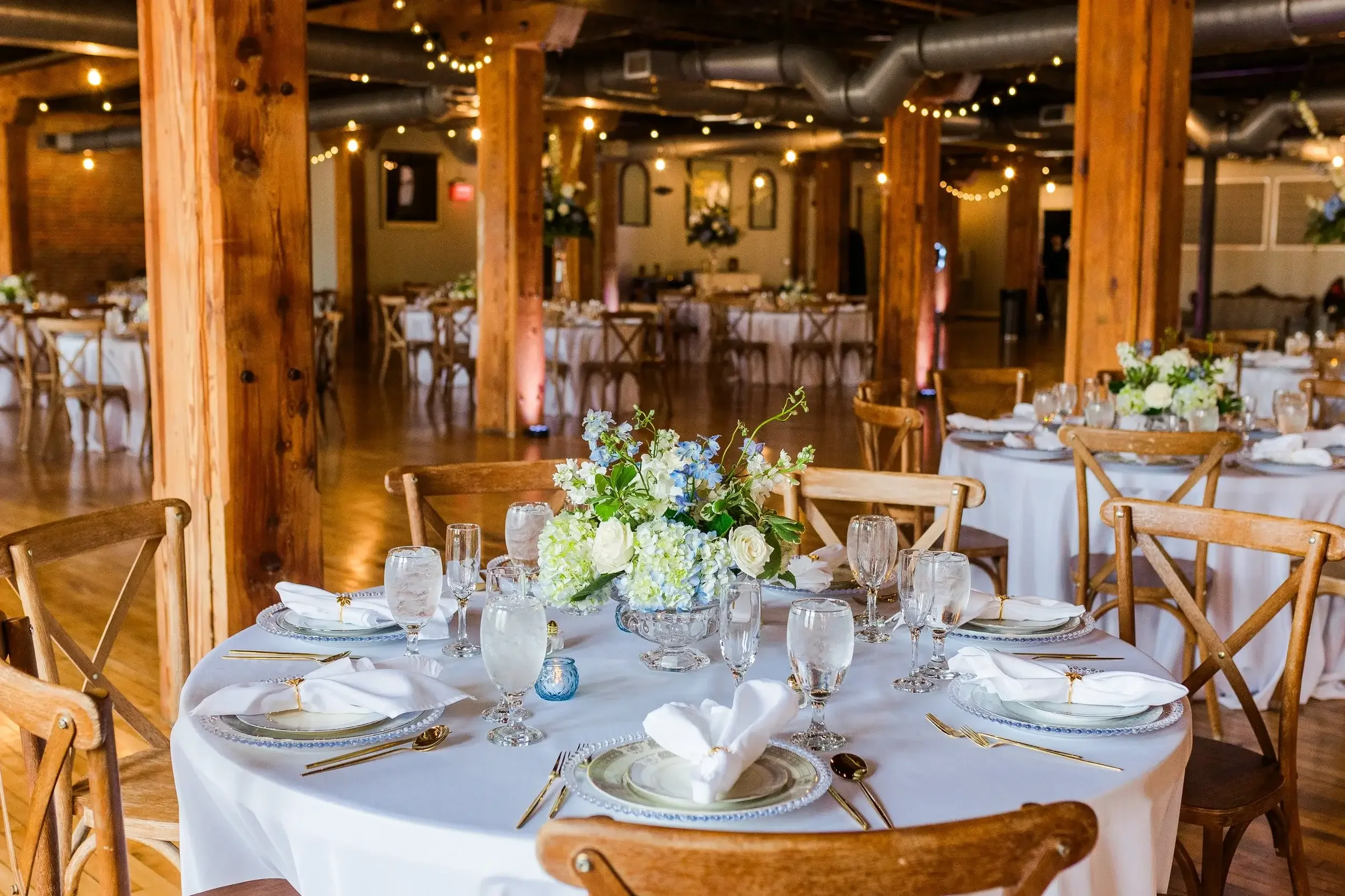 Elegant banquet hall decorated for a wedding reception with round tables, white tablecloths, floral centerpieces, crystal glassware, gold utensils, and wooden chairs. Soft lighting and string lights create a warm atmosphere.