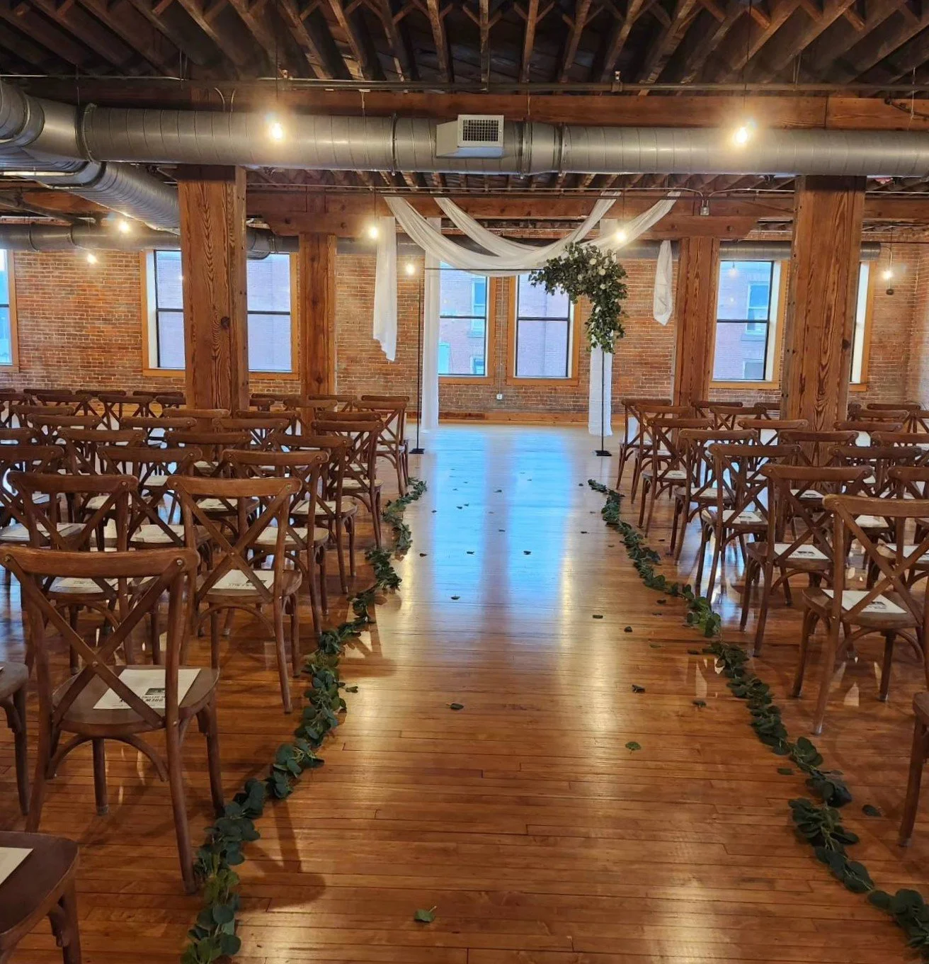 An empty indoor wedding ceremony setup with wooden chairs arranged on either side of a wooden aisle, decorated with greenery, in a brick-walled room with large windows, wooden beams, and draped fabric with hanging lights.