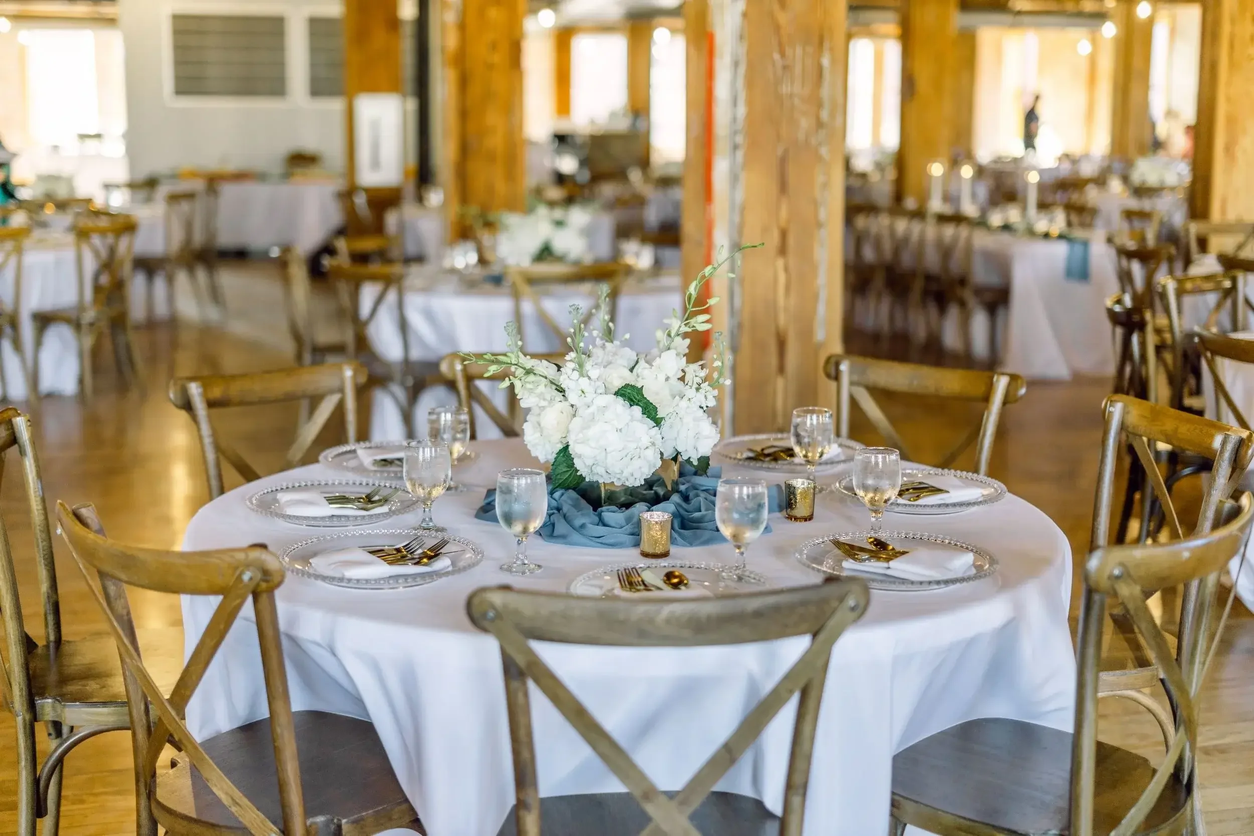 Round banquet table decorated with a white tablecloth, a floral centerpiece with white flowers, blue fabric, glassware, and gold utensils, inside a rustic wooden hall with additional decorated tables in the background.