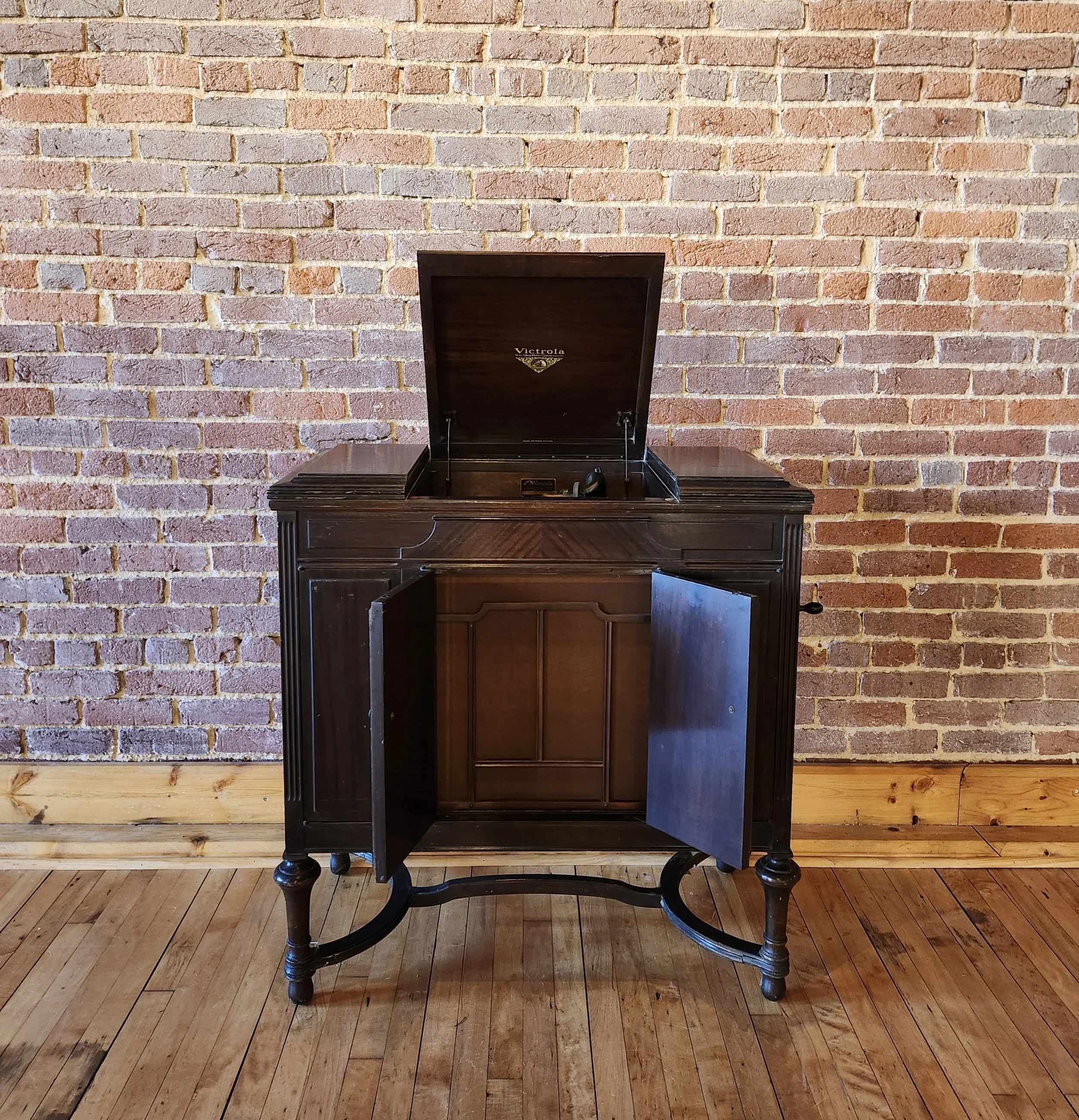 Vintage Victrola gramophone with a dark wooden cabinet, open lid, and closed lower doors, set against a brick wall background on a wooden floor.