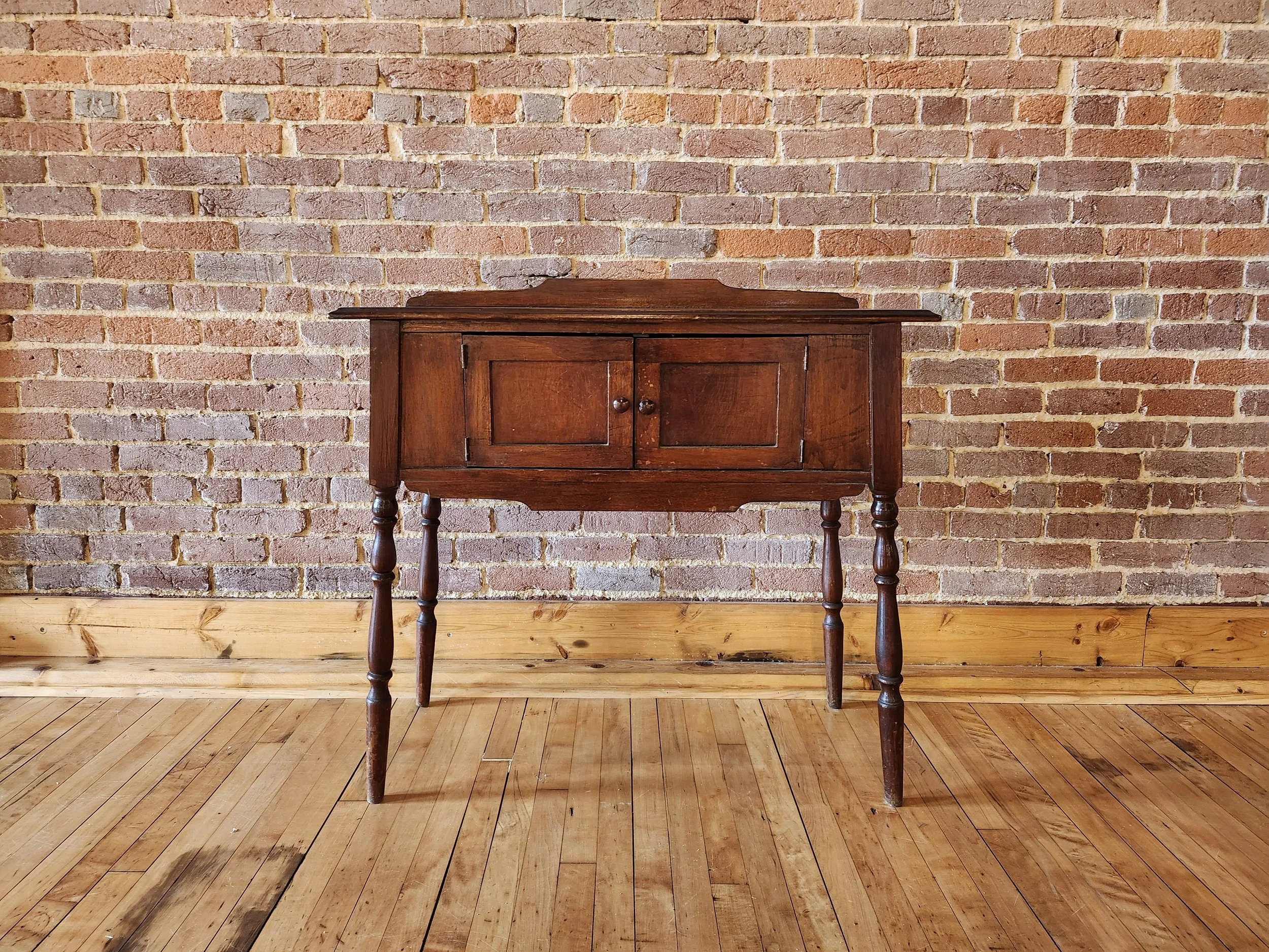 A vintage wooden table with two doors is placed against a brick wall in a room with wooden flooring.
