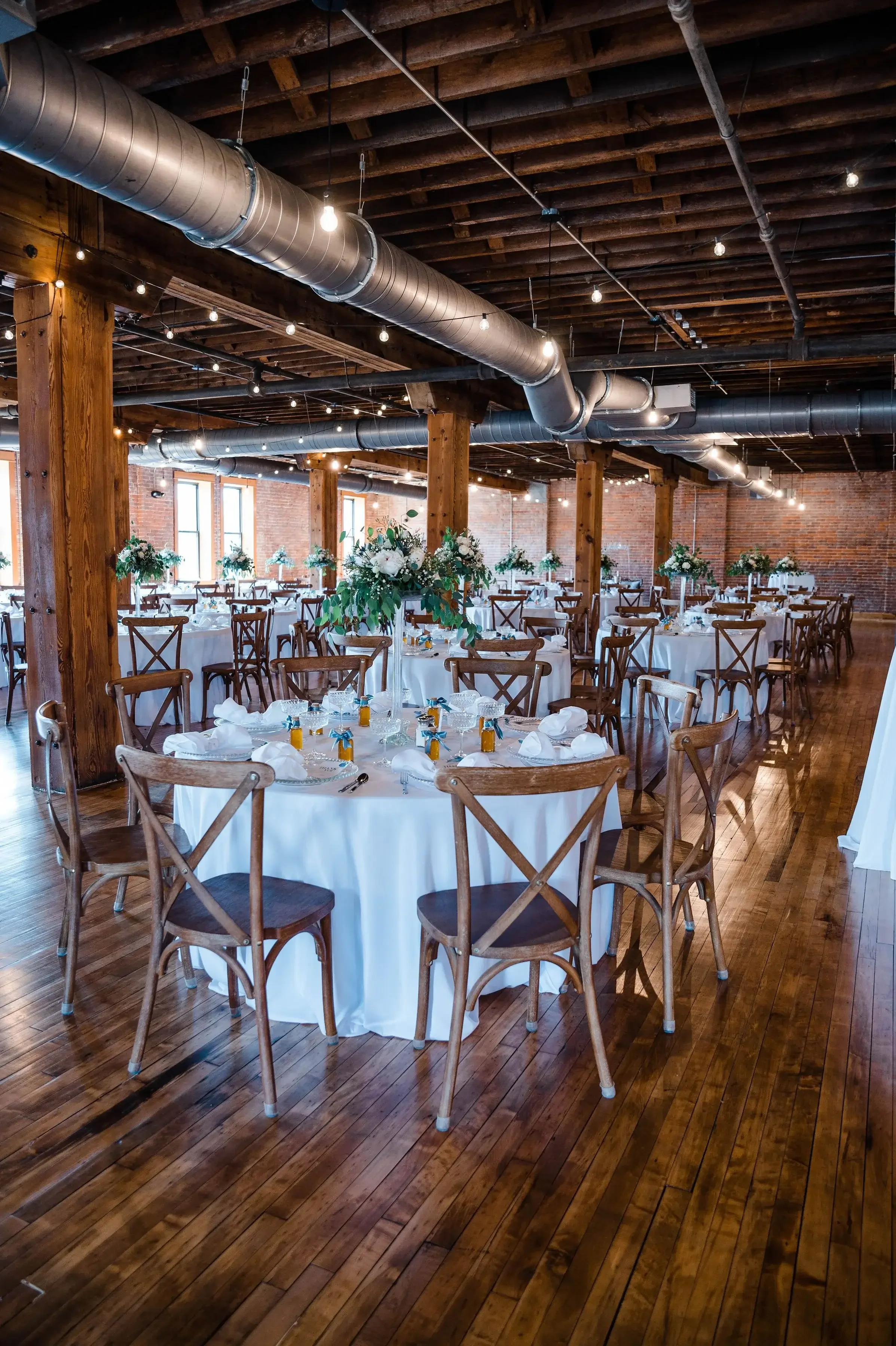 Indoor event space decorated with round tables covered in white tablecloths, set for a formal occasion, surrounded by wooden chairs with floral centerpieces, natural light coming through large windows, exposed brick walls, and industrial ceiling pipe