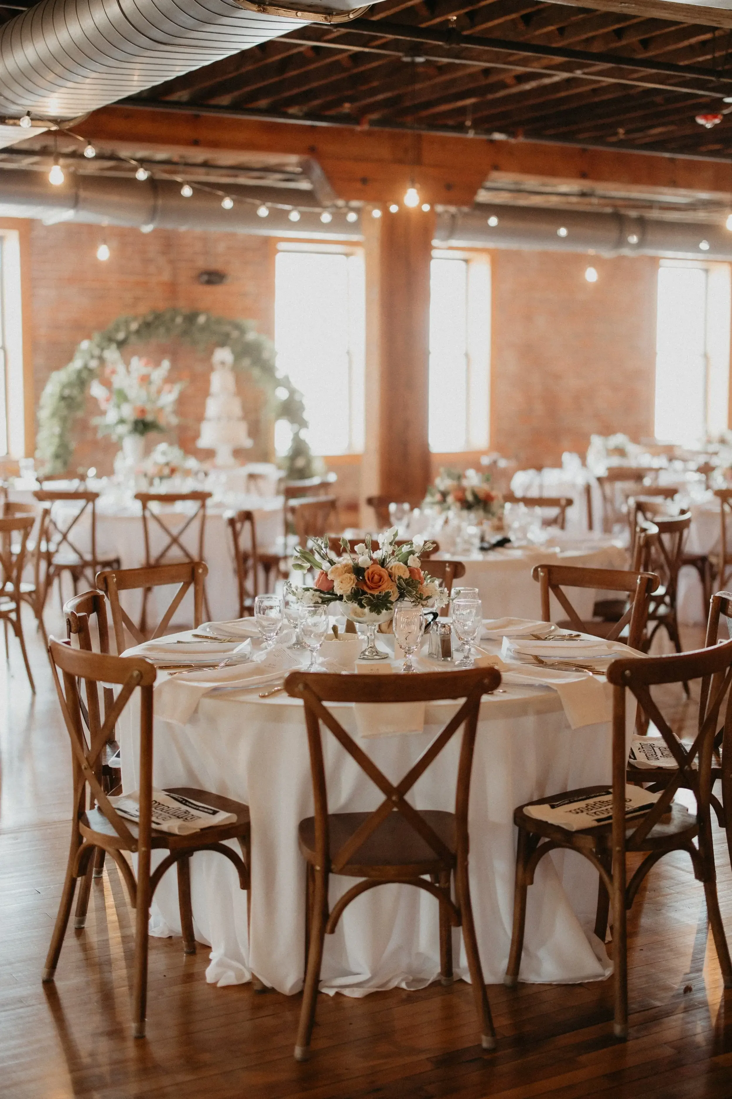 Elegant wedding reception setup with round table covered in white tablecloth, adorned with floral centerpiece and fine glassware in a rustic wooden venue with string lights and large windows.