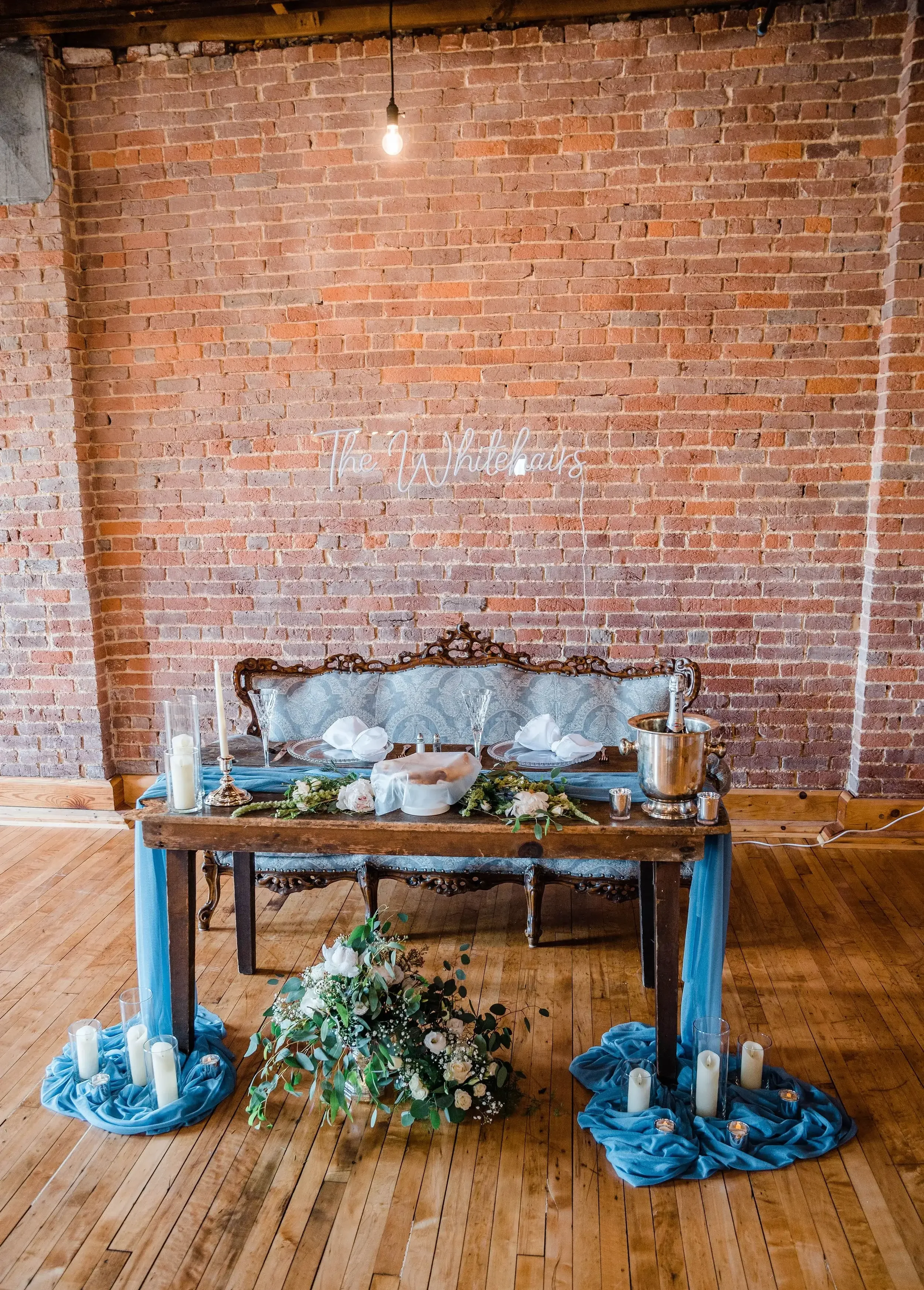 Wedding reception table setup with floral arrangements, candles, and a blue draped tablecloth, against a brick wall with a neon sign that reads 'The Whitehairs'.