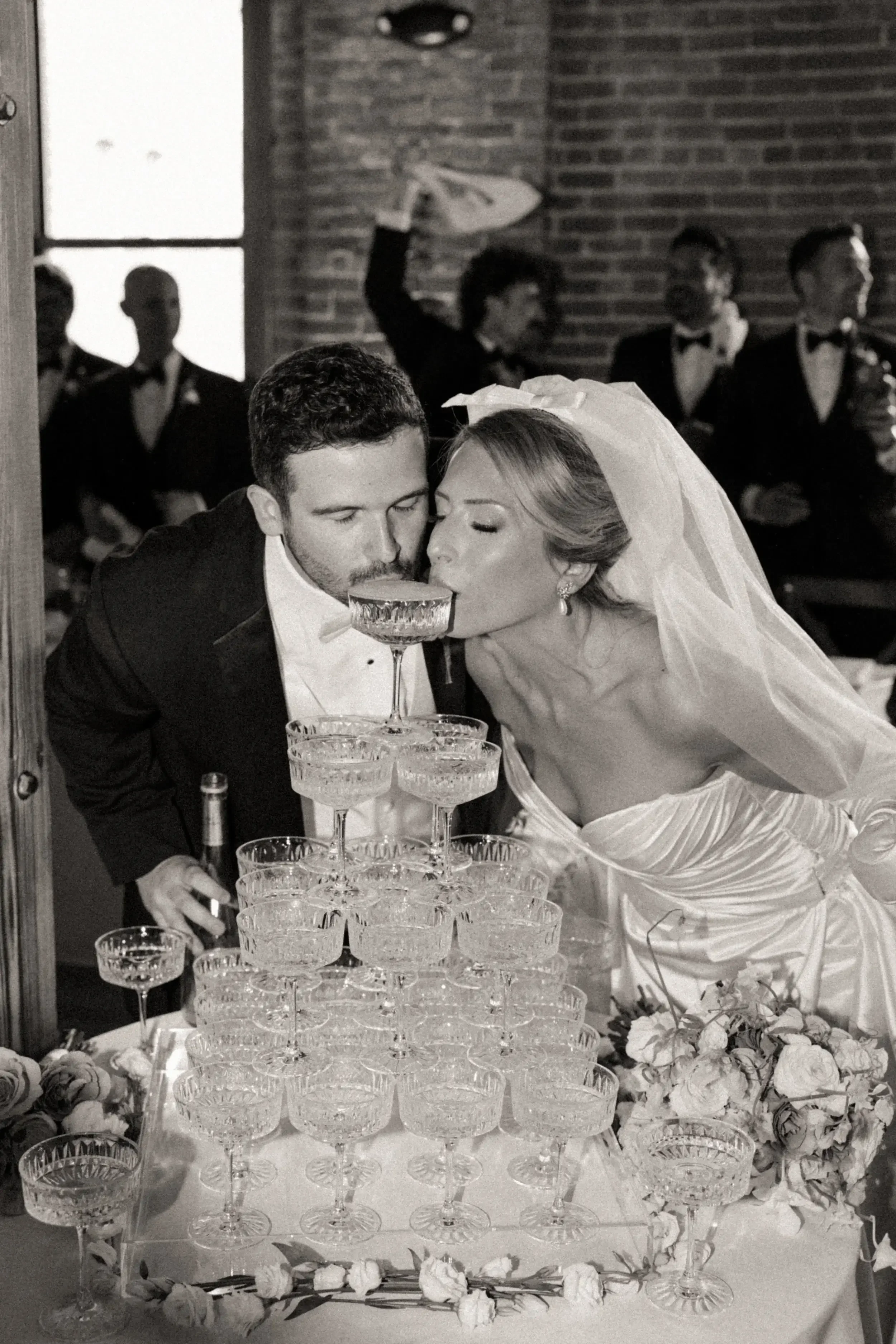 A black and white photo of a wedding reception where a bride and groom are taking a shot together from a champagne tower. The bride is wearing a wedding dress and veil, and the groom is in a tuxedo. In the background, a band with musicians dressed in