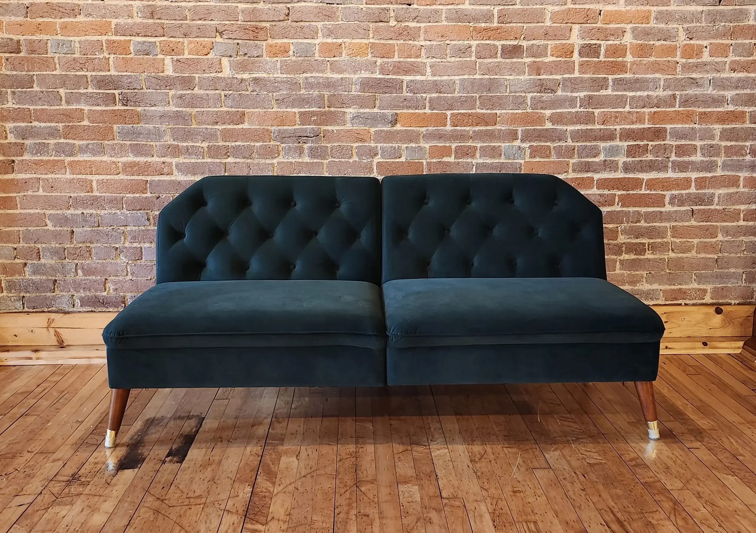 A vintage-style dark green velvet sofa with wooden legs and brass tips, positioned against a brick wall on a wooden floor.
