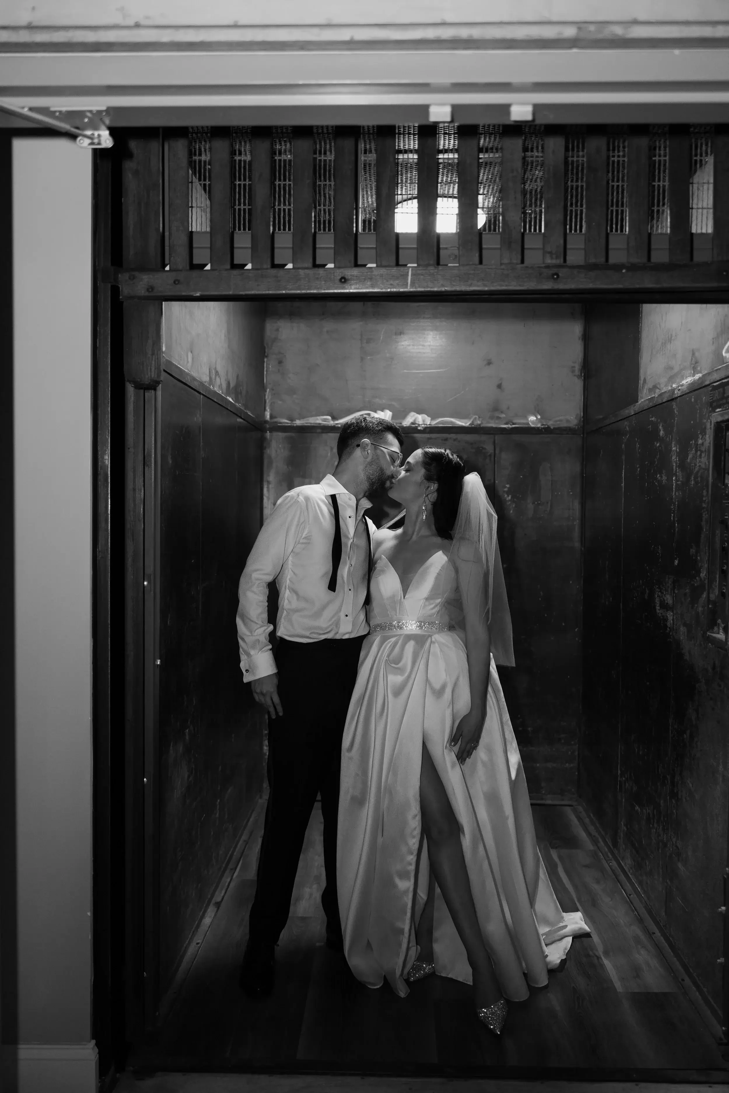 A black and white photo of a newlywed couple kissing in an elevator or small enclosed space, with wooden walls and a decorative light overhead.