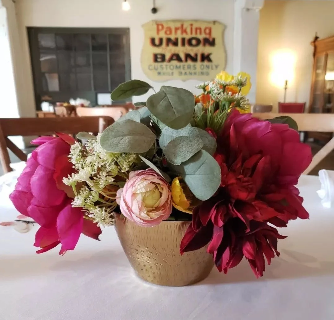A floral arrangement with pink, red, yellow, and white flowers in a gold-colored vase on a white table, with a vintage-style sign reading 'Parking Union Bank Customers Only' in the background.