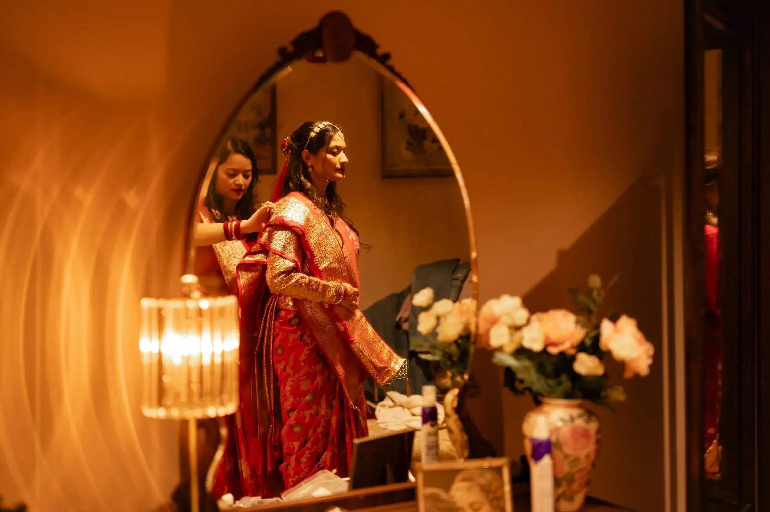Two women in traditional red and gold dresses, one helping the other, are seen through a mirror in a warmly lit room with flowers and personal items on a dresser.