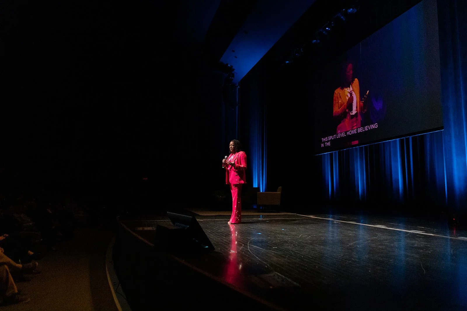 A woman in a pink suit is speaking on stage at a conference, holding a microphone. There is a large screen behind her showing her image and some text. Sophia Fifner Keynote Speaker