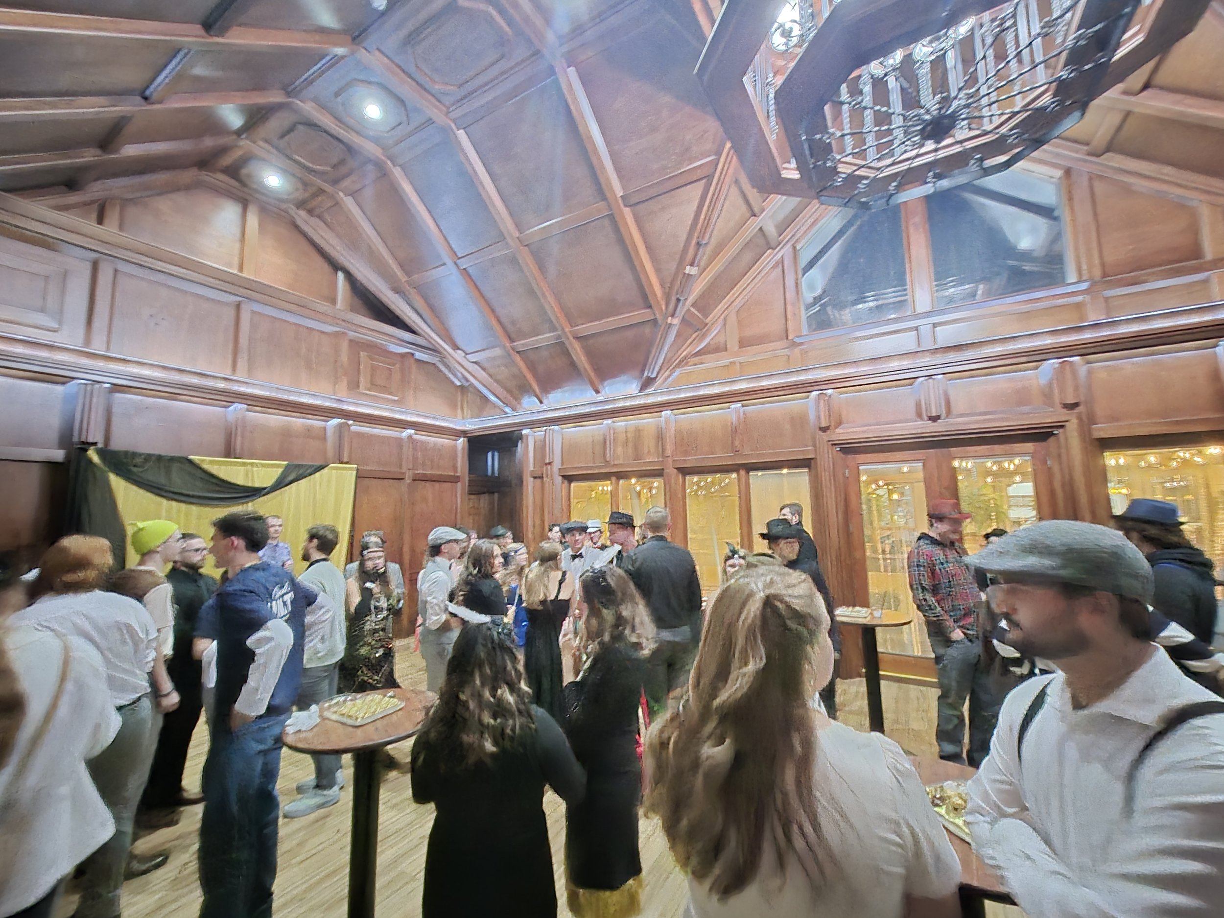 People gathered at an indoor social event in a wood-paneled room with high ceiling and large windows, some holding drinks and conversation.