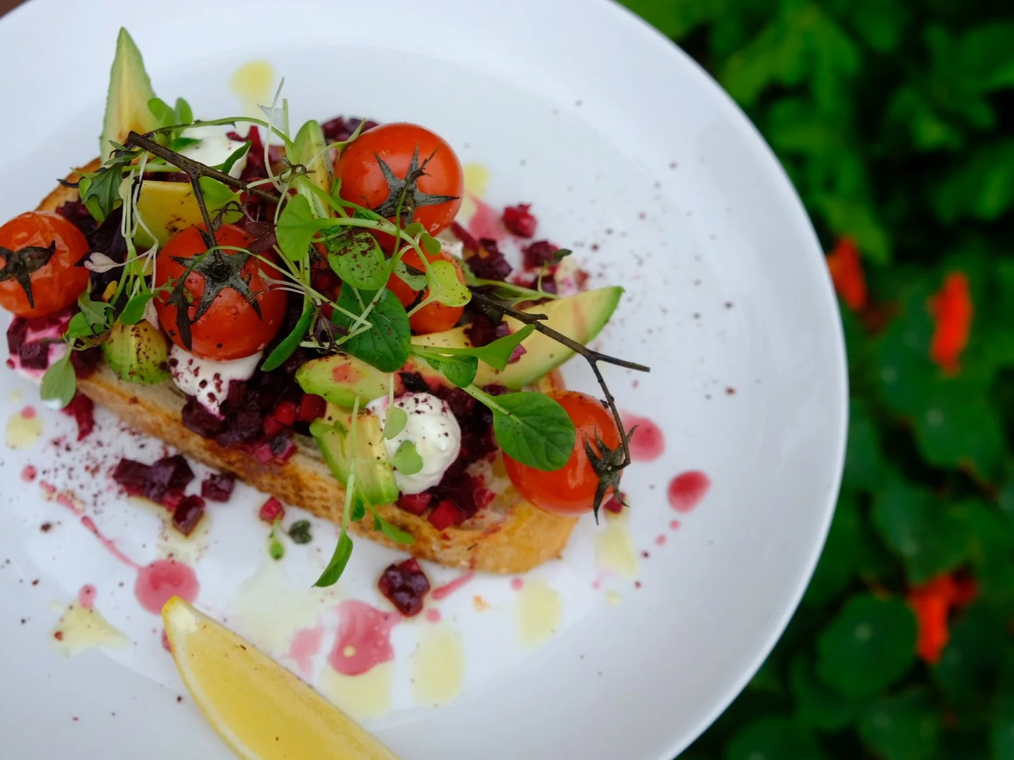 Vegetable toast topped with cherry tomatoes, avocado slices, microgreens, and drizzled with oil and balsamic vinegar on a white plate with lemon wedge, set outdoors
