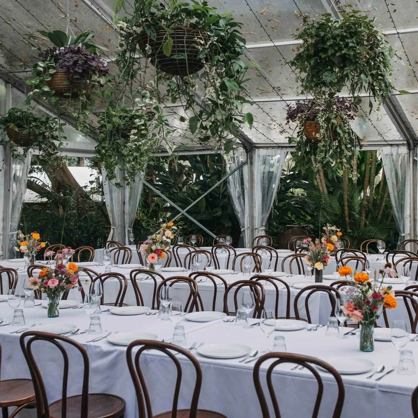 Elegant indoor event setup with long white-draped tables, floral centerpieces, and wooden chairs, surrounded by lush greenery and hanging plants in a glass-walled venue.