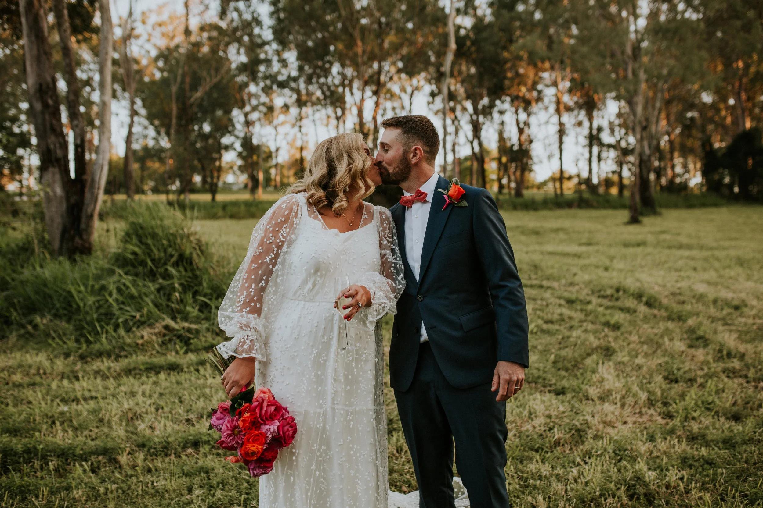 A bride and groom sharing a kiss outdoors during their wedding, with the bride holding a bouquet of pink and red flowers and the groom wearing a dark suit with a pink bow tie and a red boutonniere, in a grassy area with trees in the background at sunset.