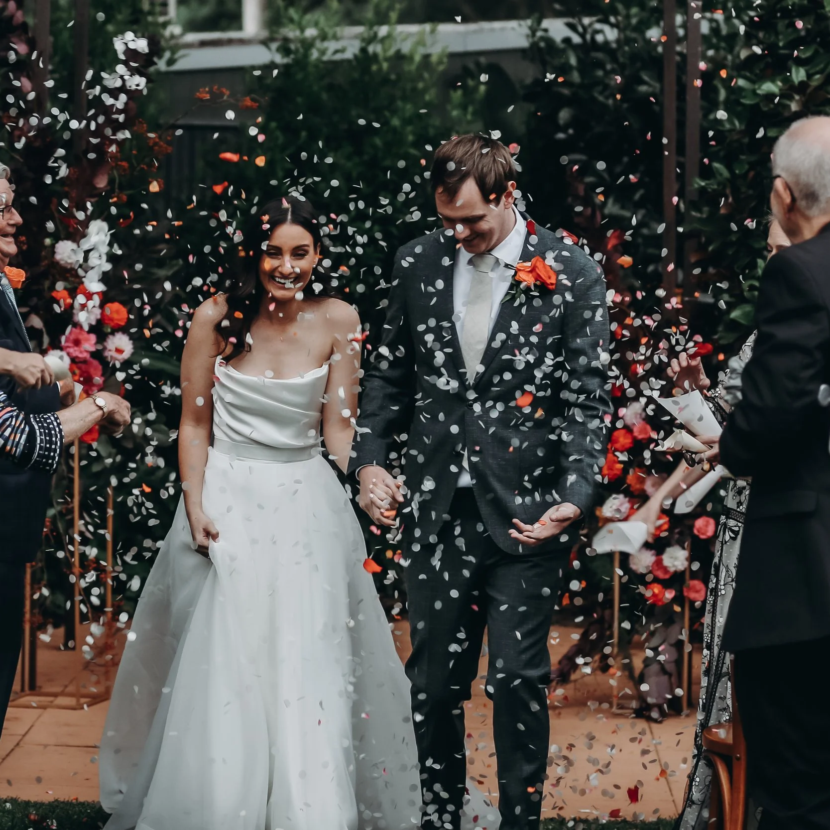 A bride and groom walking hand in hand at their wedding reception, surrounded by guests and showered with confetti in an outdoor garden setting.