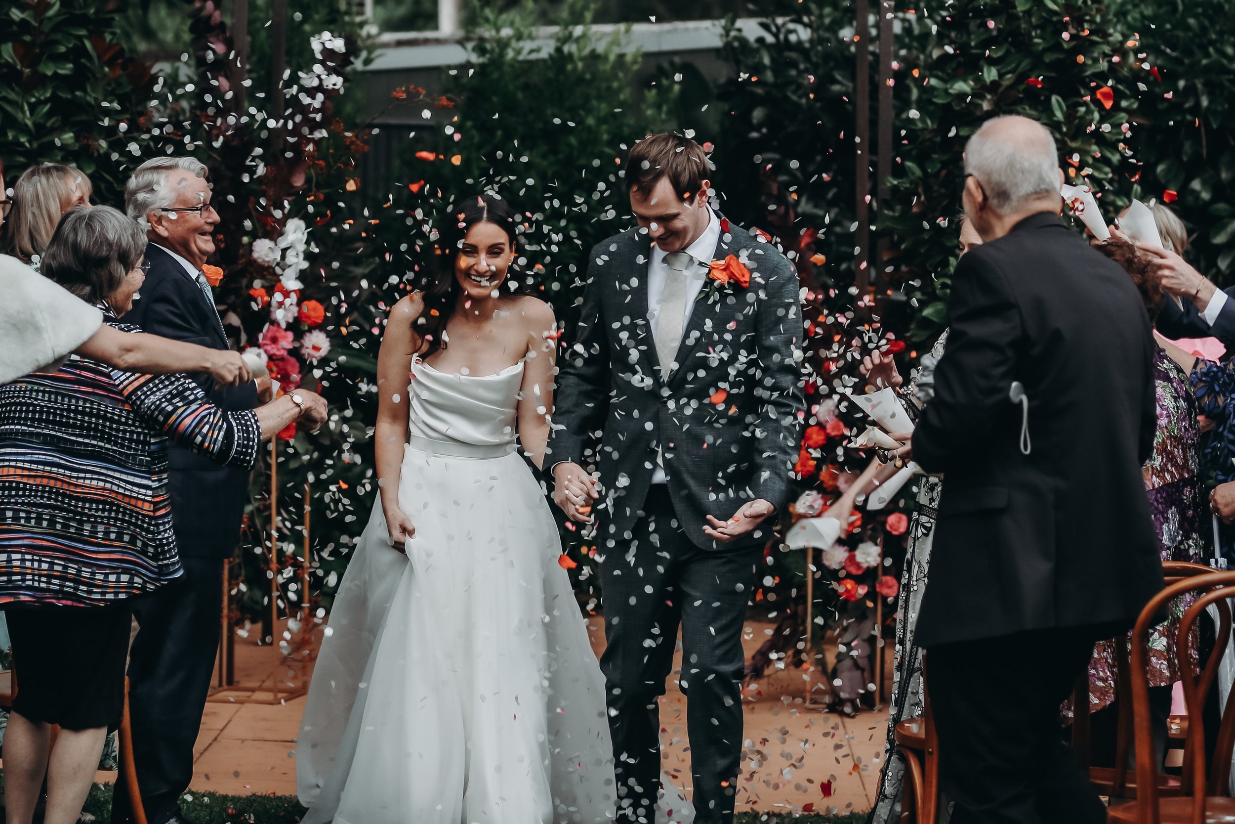 A newly married couple holding hands and smiling as confetti is thrown around them during their wedding celebration outdoors, with guests on both sides celebrating.