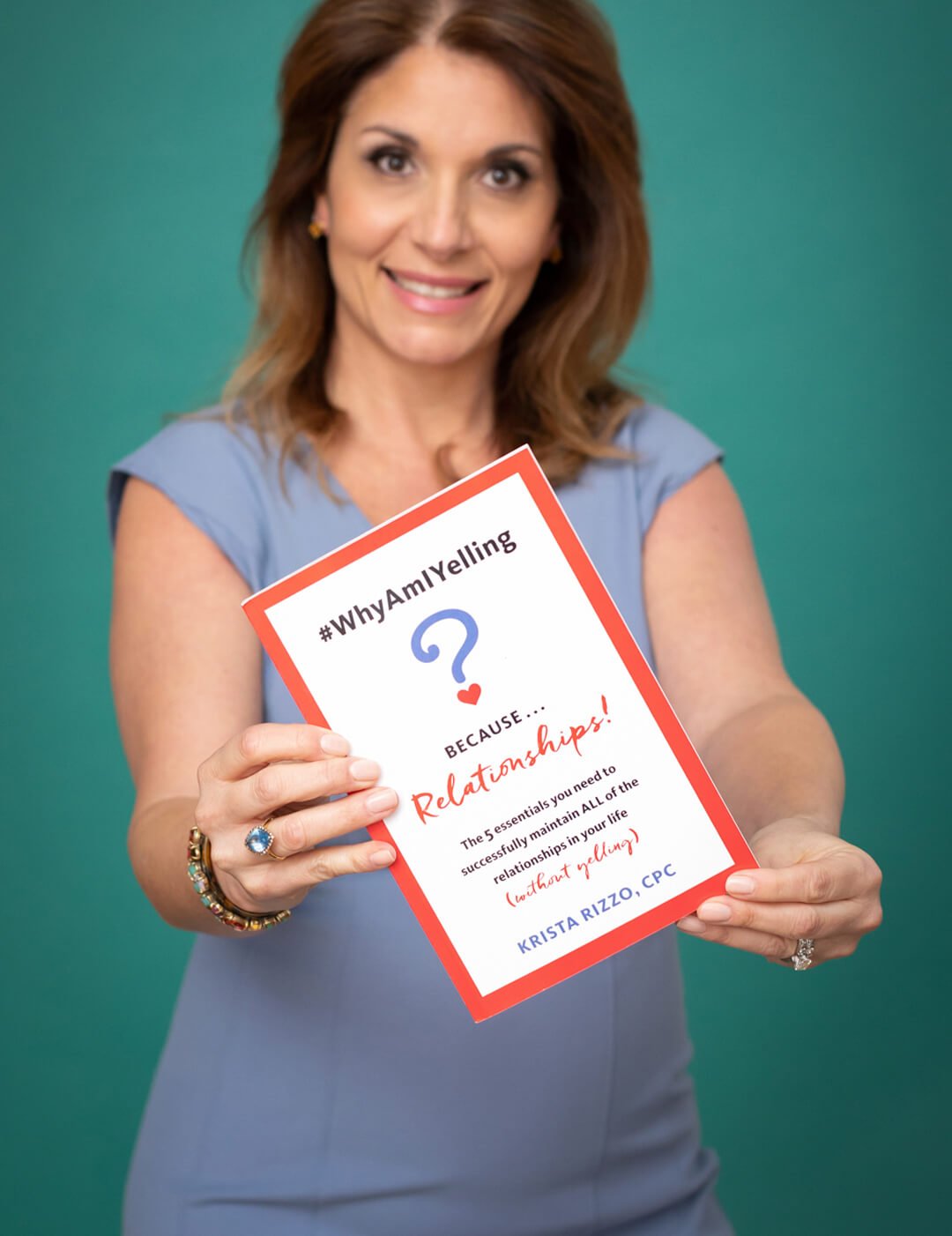 Krista Rizzo holding her book "Why Am I Yelling?!" while smiling. Krista is a white woman, wearing a light blue dress and jewelry. She has orange mid-length hair.