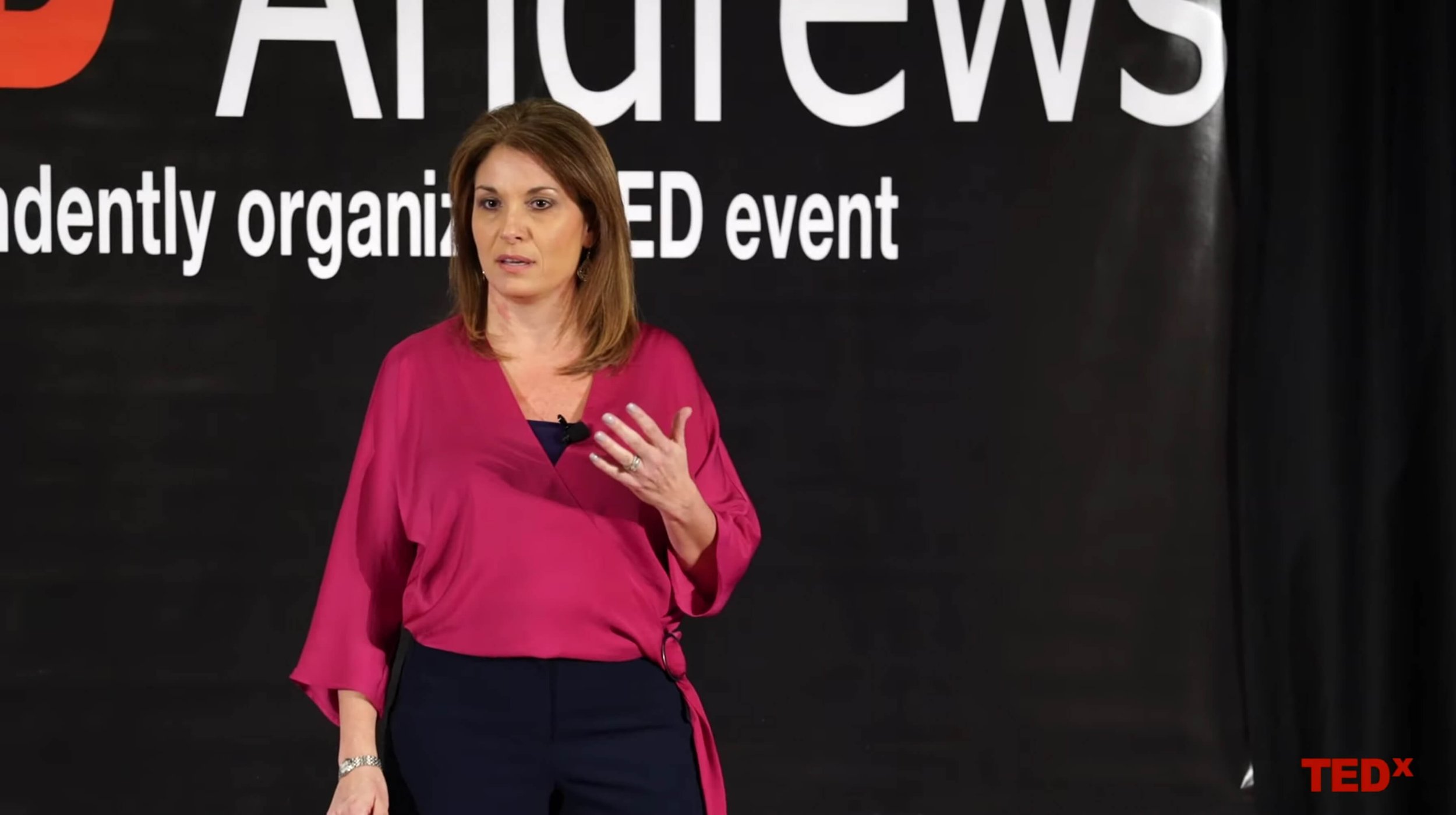 Krista Rizzo on the stage of TEDx while she delivers her speech. Krista is a white woman, wearing a fucsia top and blue pants. She has orange mid-length hair.
