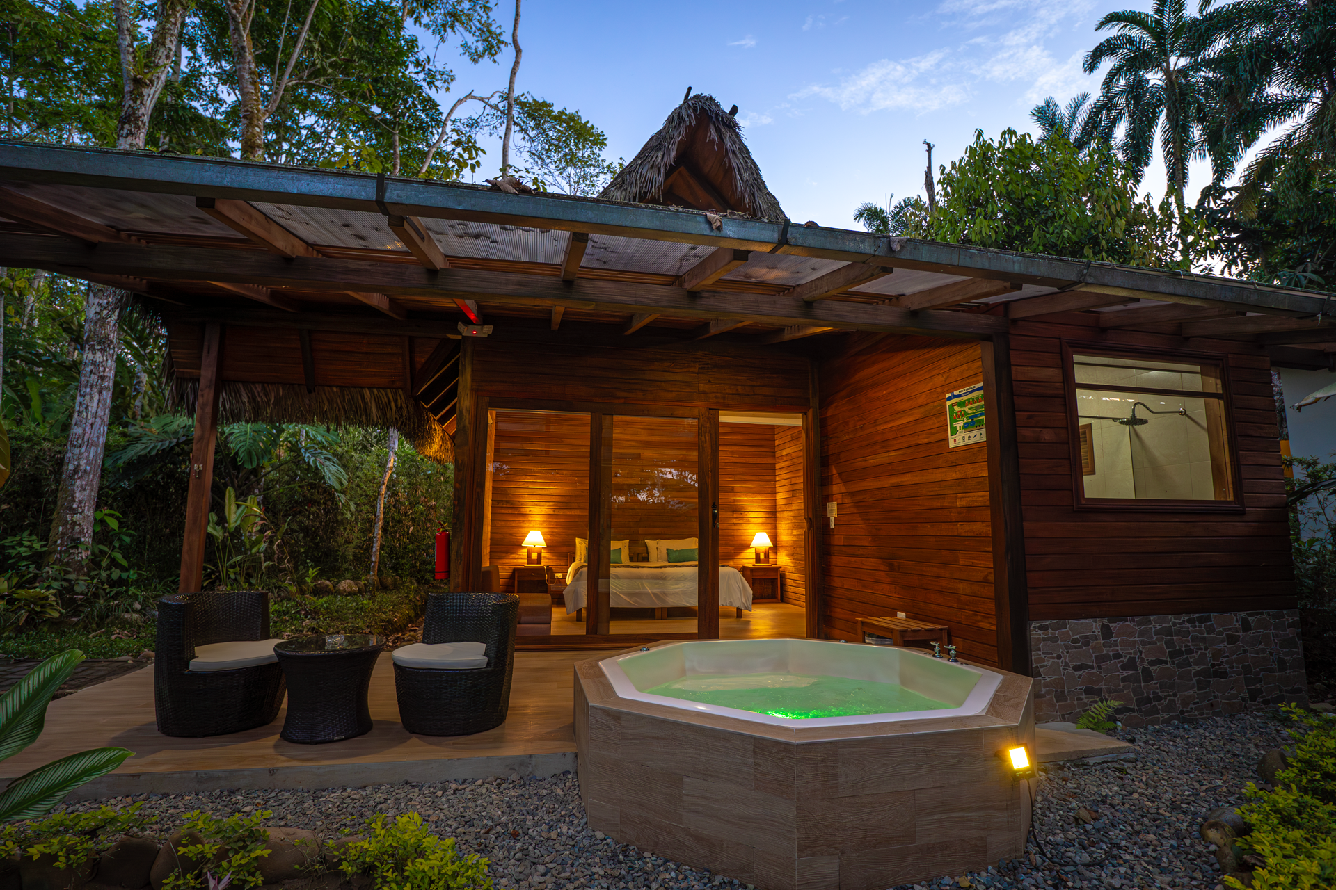 Cabaña de madera rústica con jacuzzi exterior iluminado, rodeada de vegetación tropical, muebles de exterior y una habitación visible con luces cálidas encendidas en el interior.
