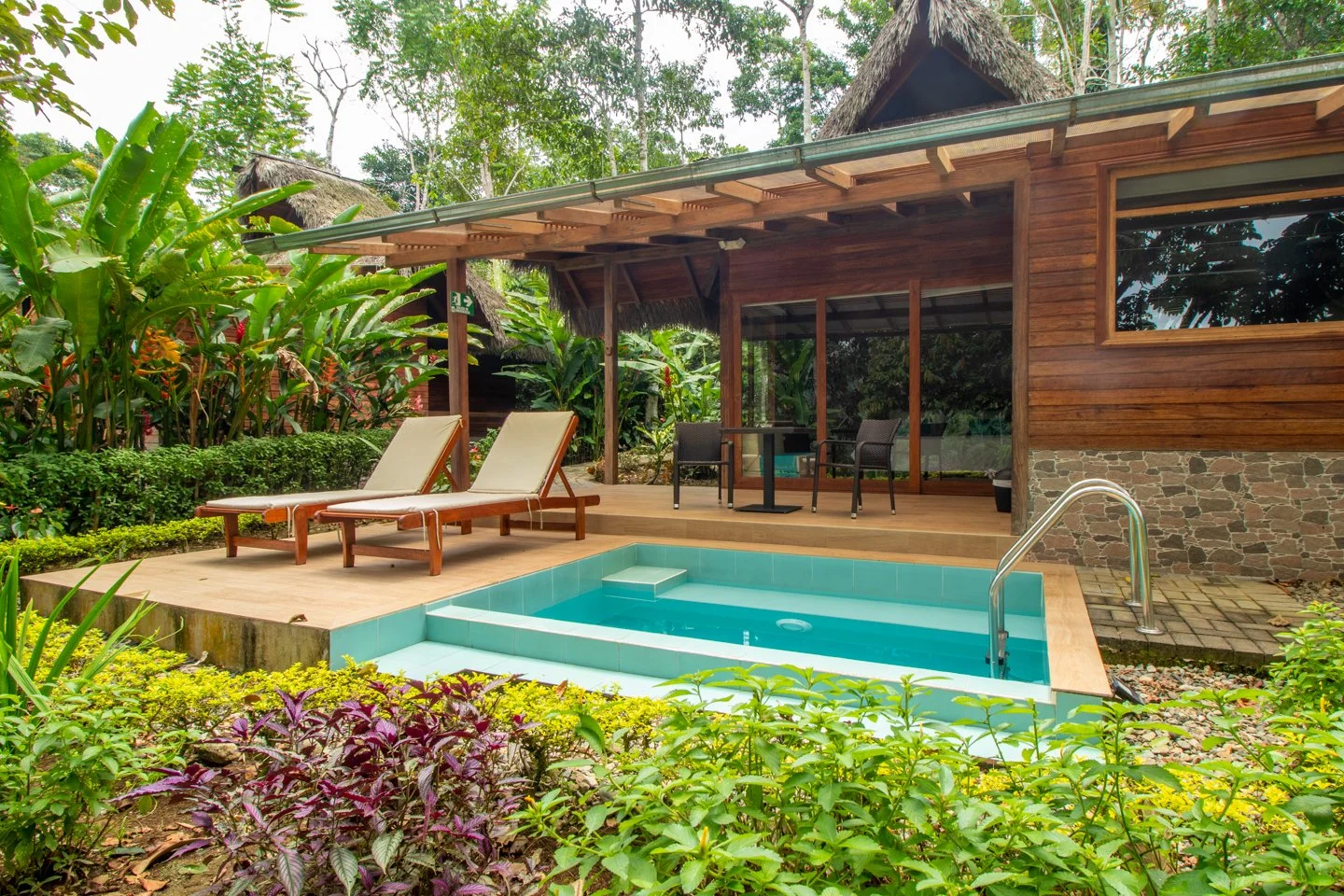 Cabaña de madera con piscina pequeña rodeada de vegetación tropical, con dos tumbonas en una terraza de madera.