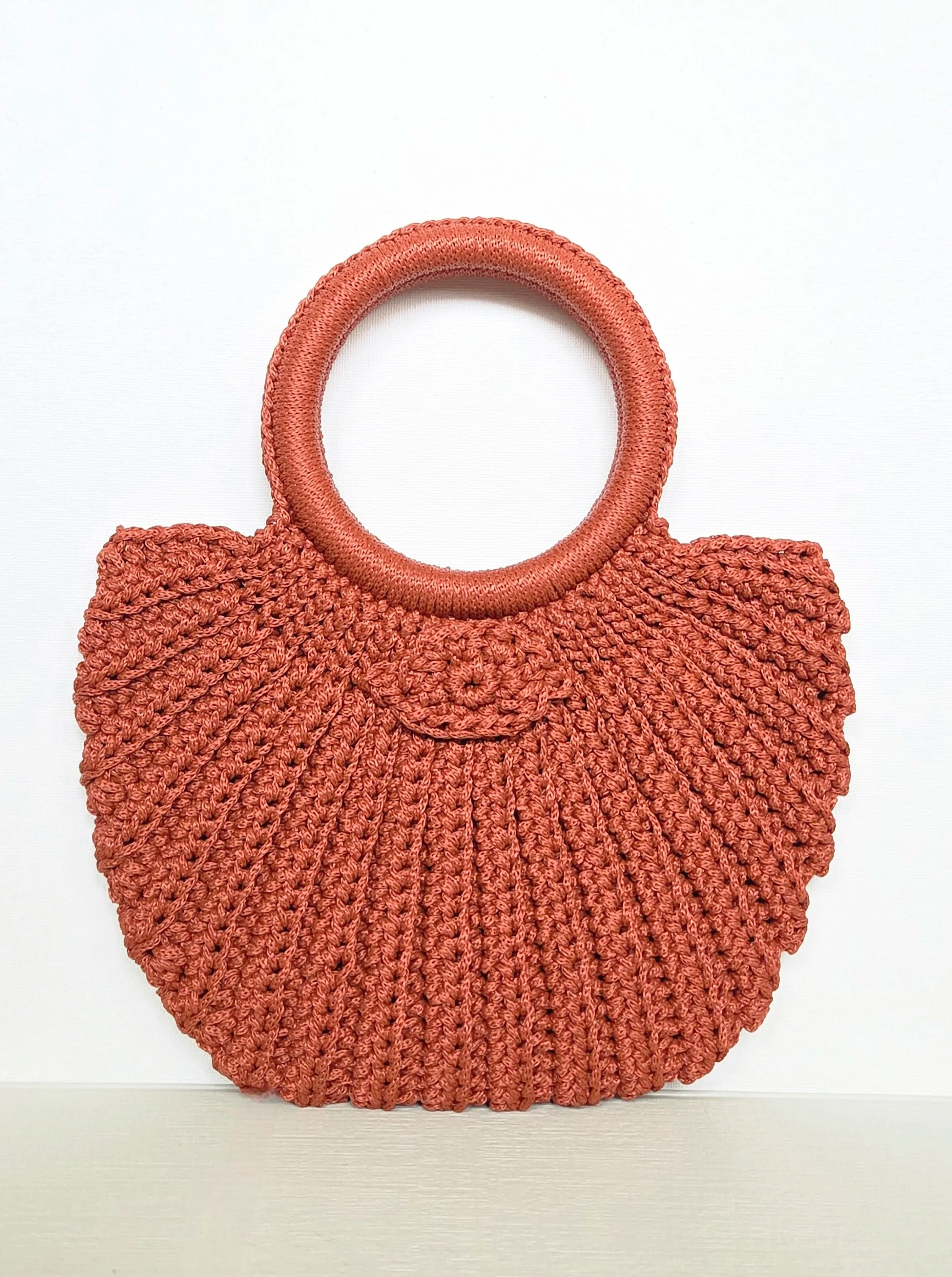 Coral-colored, knitted handbag with circular handle and woven texture, displayed against a plain white background.