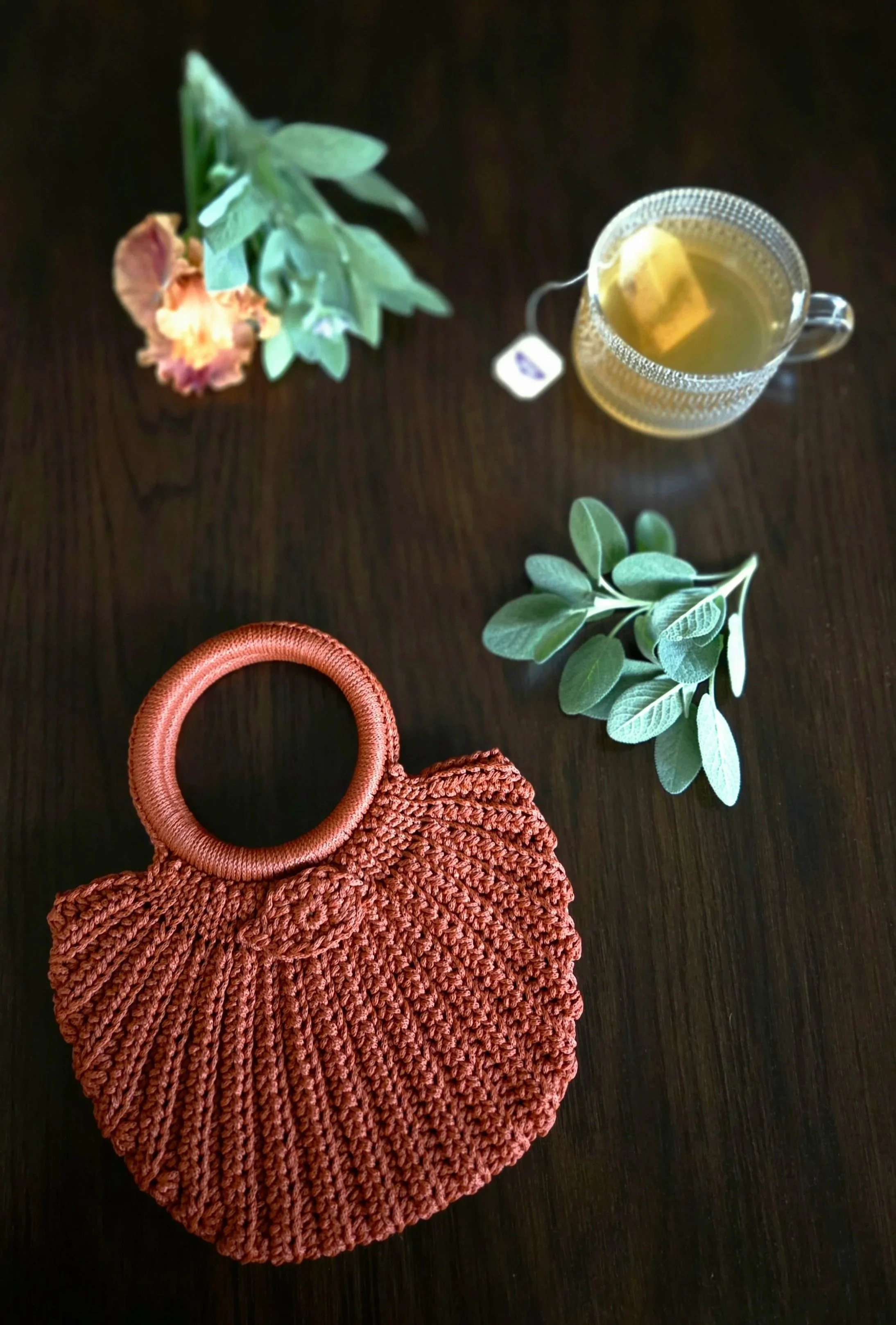 A red woven purse with a circular handle next to a green leafy branch, a glass cup of tea with a tea bag, a sprig of sage, and a peach-colored flower on a dark wooden surface.