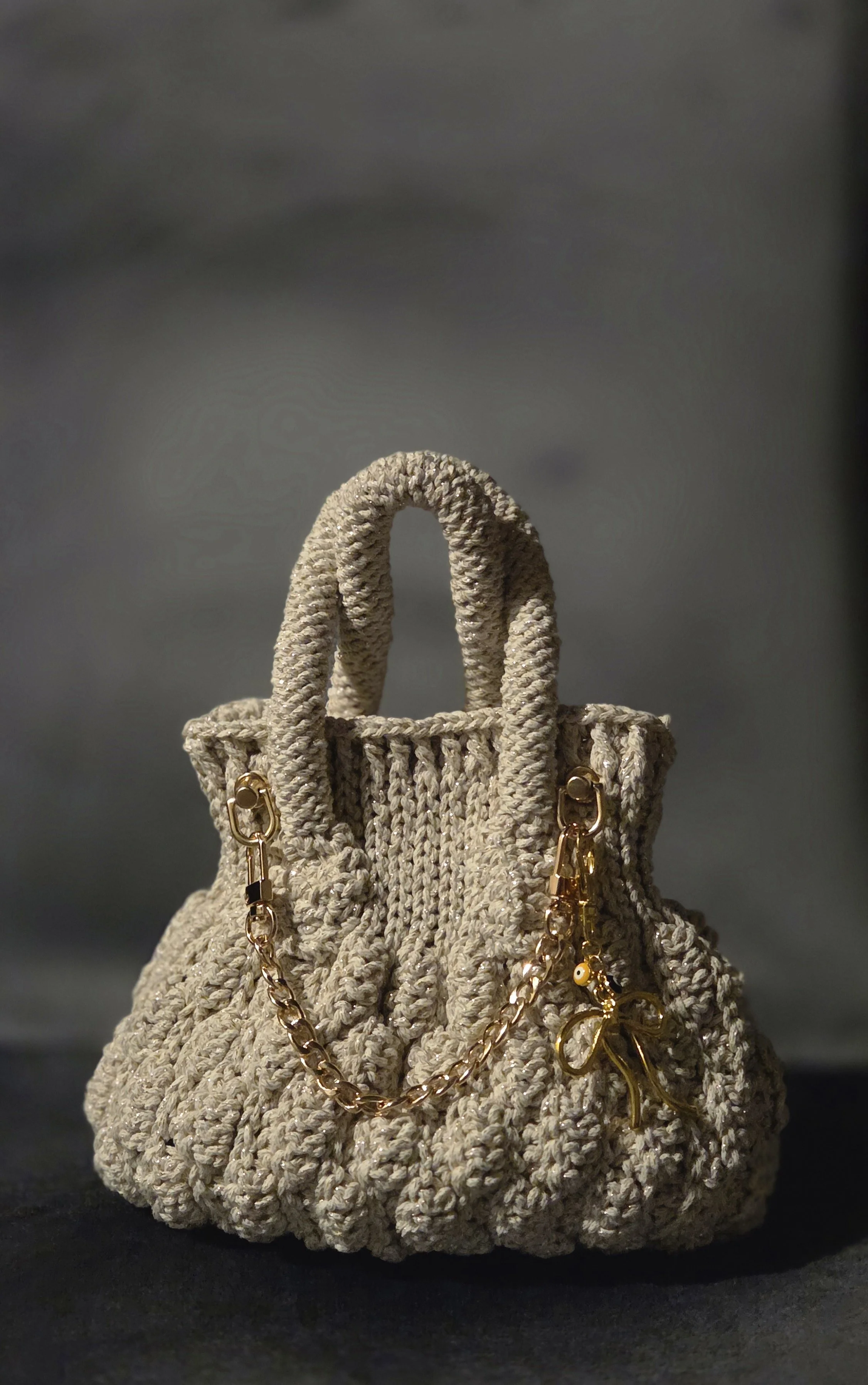 A beige woven handbag with gold chain straps and decorative charms, placed against a dark background.