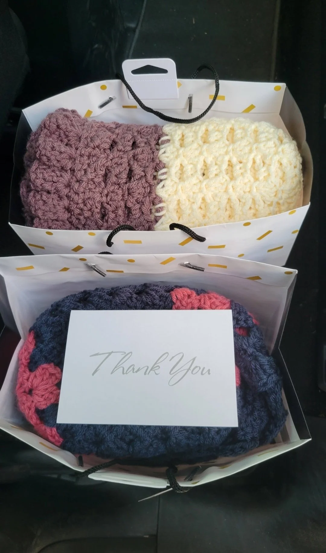 Two gift bags containing crochet hats, one with pink and purple yarn, and the other with black, pink, and purple yarn, with a white card reading 'Thank You' placed on top of the second bag.