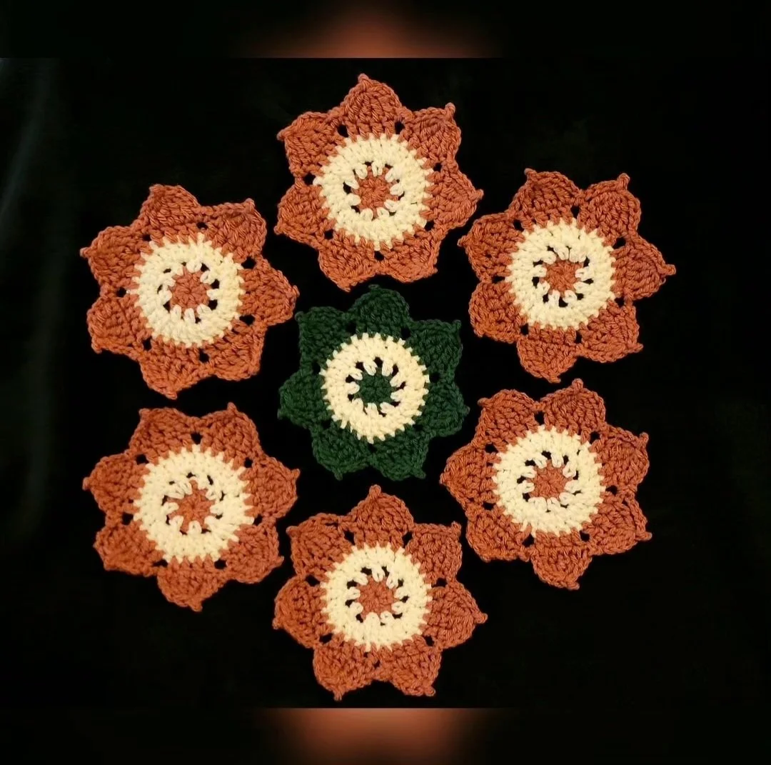 Six crocheted flowers with a central white circle, surrounded by orange petals, and one green flower in the middle.