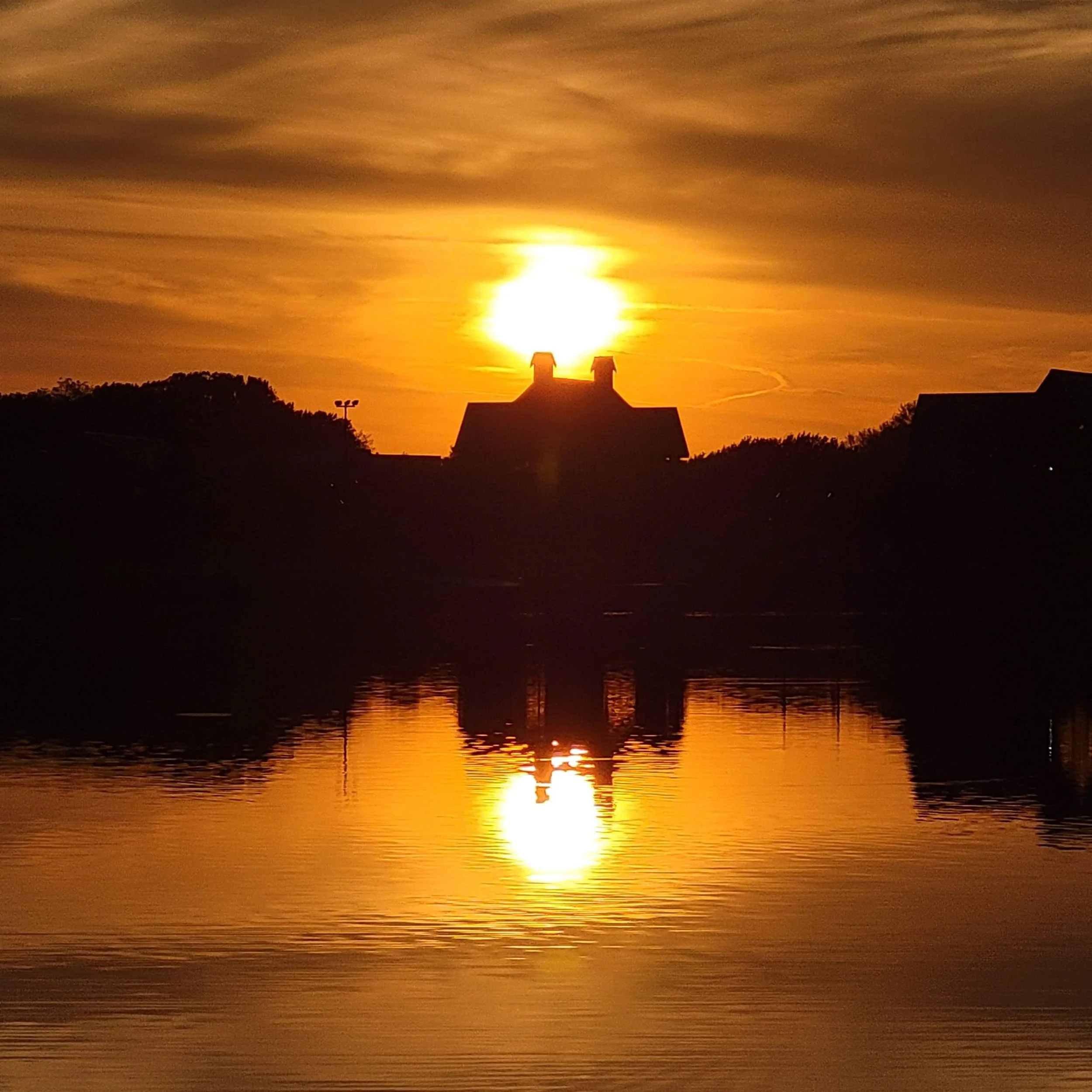Sun setting over a body of water with orange and yellow hues, silhouettes of buildings and trees in the foreground.