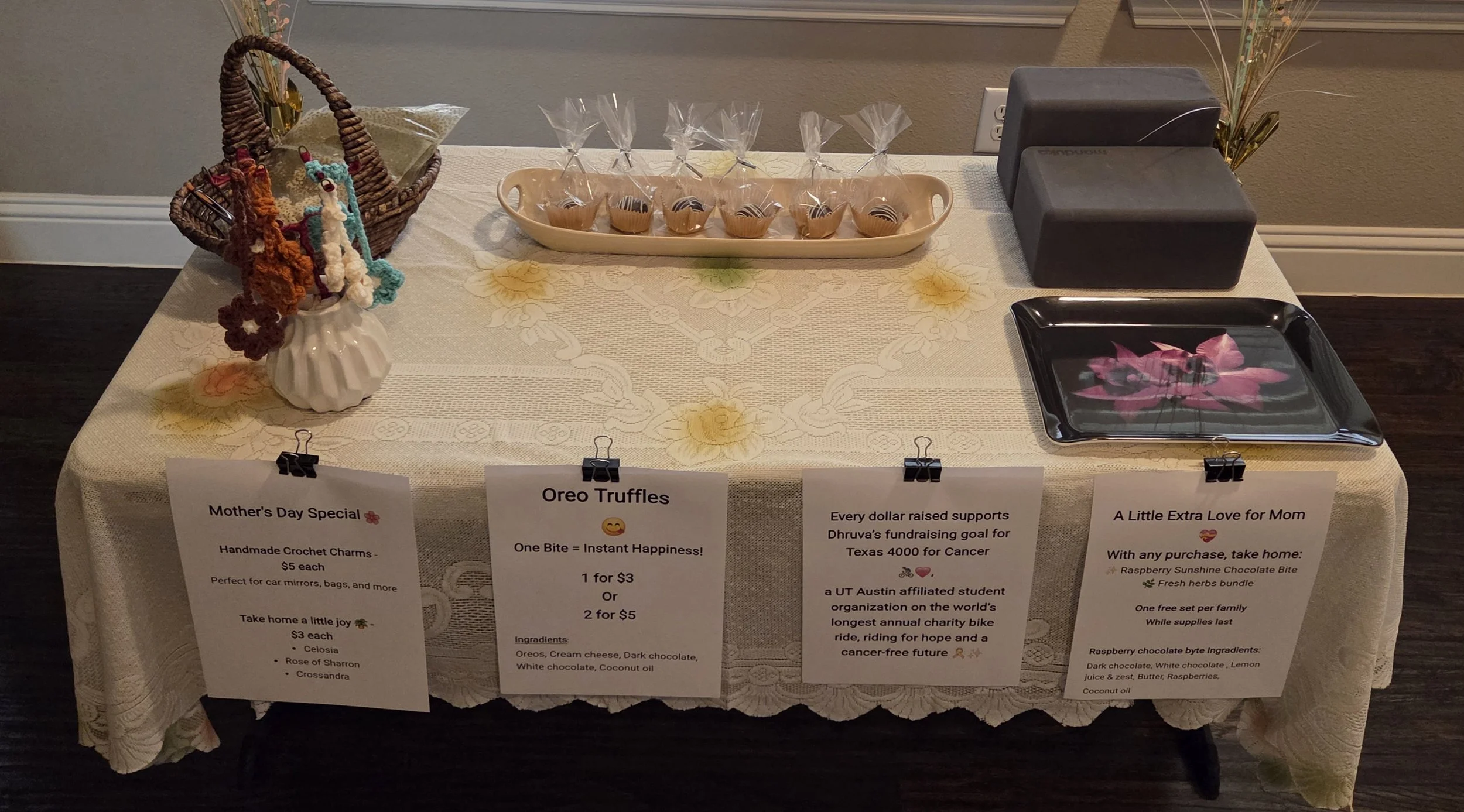 A table with a cloth cover displaying various items for sale including handmade crochet charms, Oreo truffles, and a black tray with a pink flower design. There are signs below listing prices and descriptions, and a basket with handmade crochet charms. The setting appears to be at a fundraising event, possibly for Mother's Day.