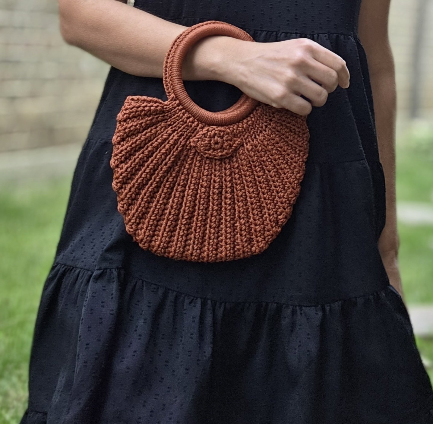 Close-up of a person holding a small, round, orange woven handbag with a loop handle. The person is wearing a black dress with a textured pattern.