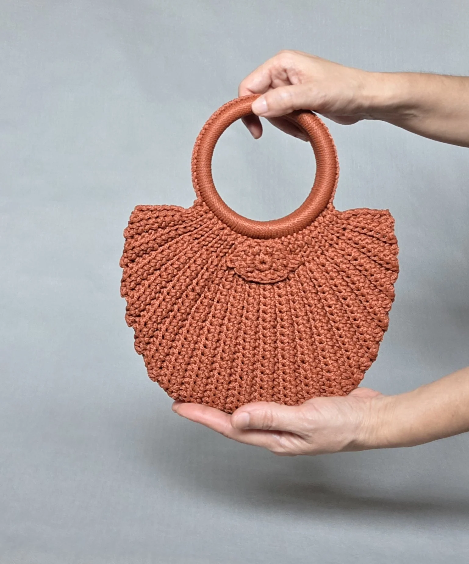 A person holding a coral-colored, woven handbag with a round handle and a floral embellishment on the front.