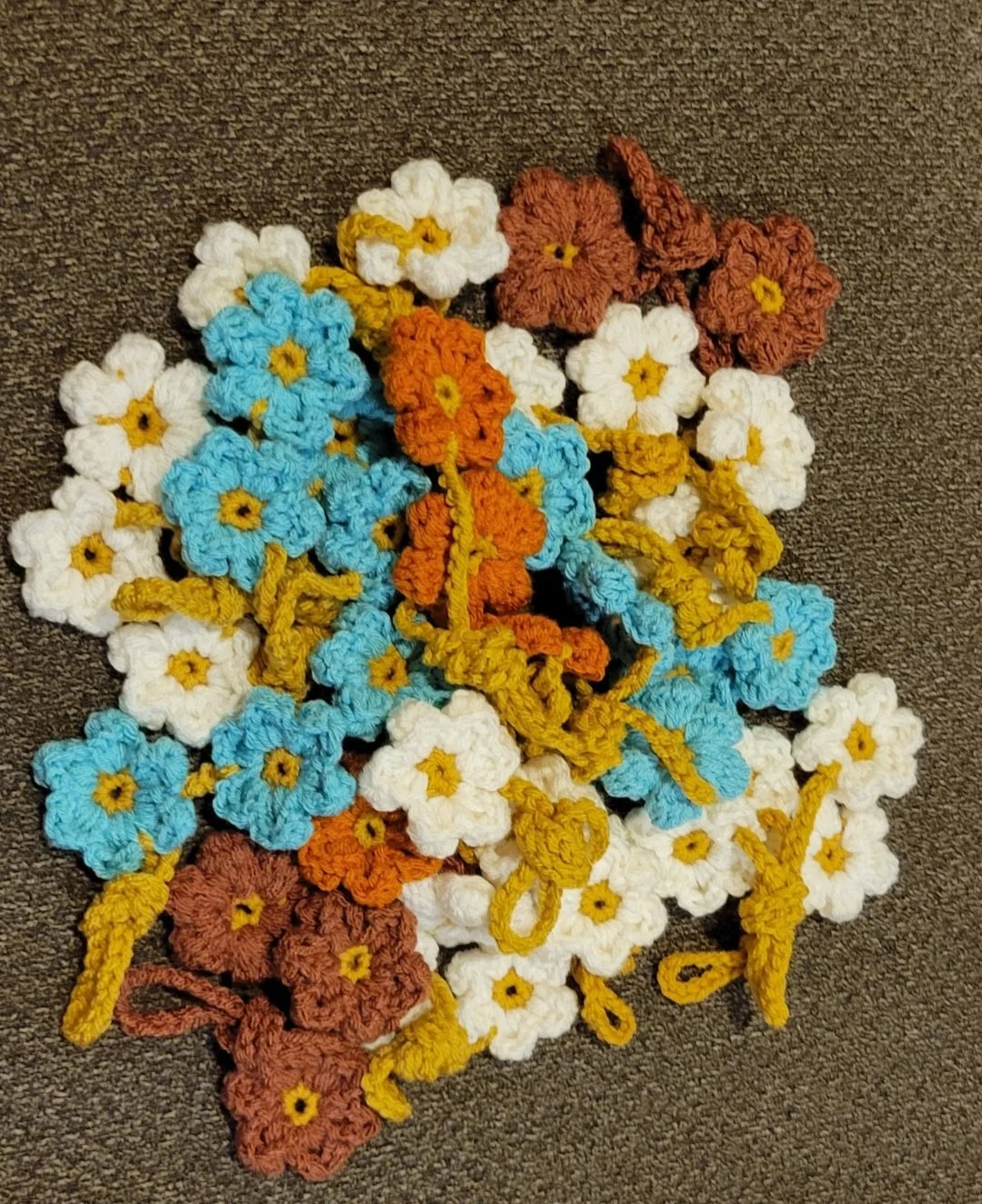 A collection of colorful crocheted flowers in white, blue, orange, and brown, arranged on a dark fabric surface.