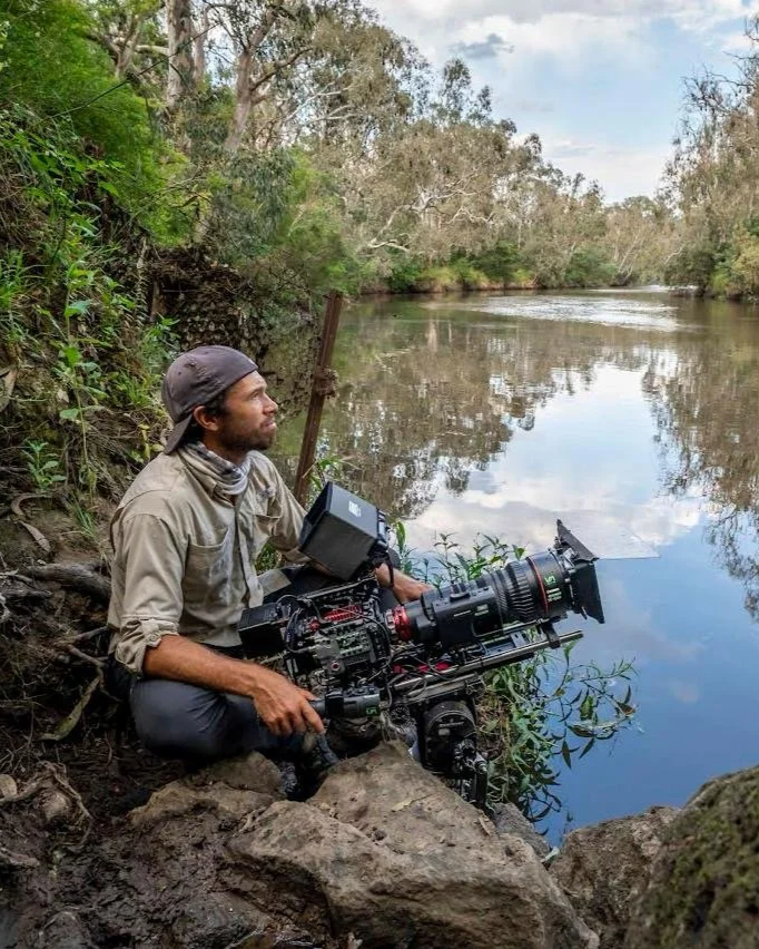A man operating a professional film camera by a river, surrounded by trees and natural scenery.