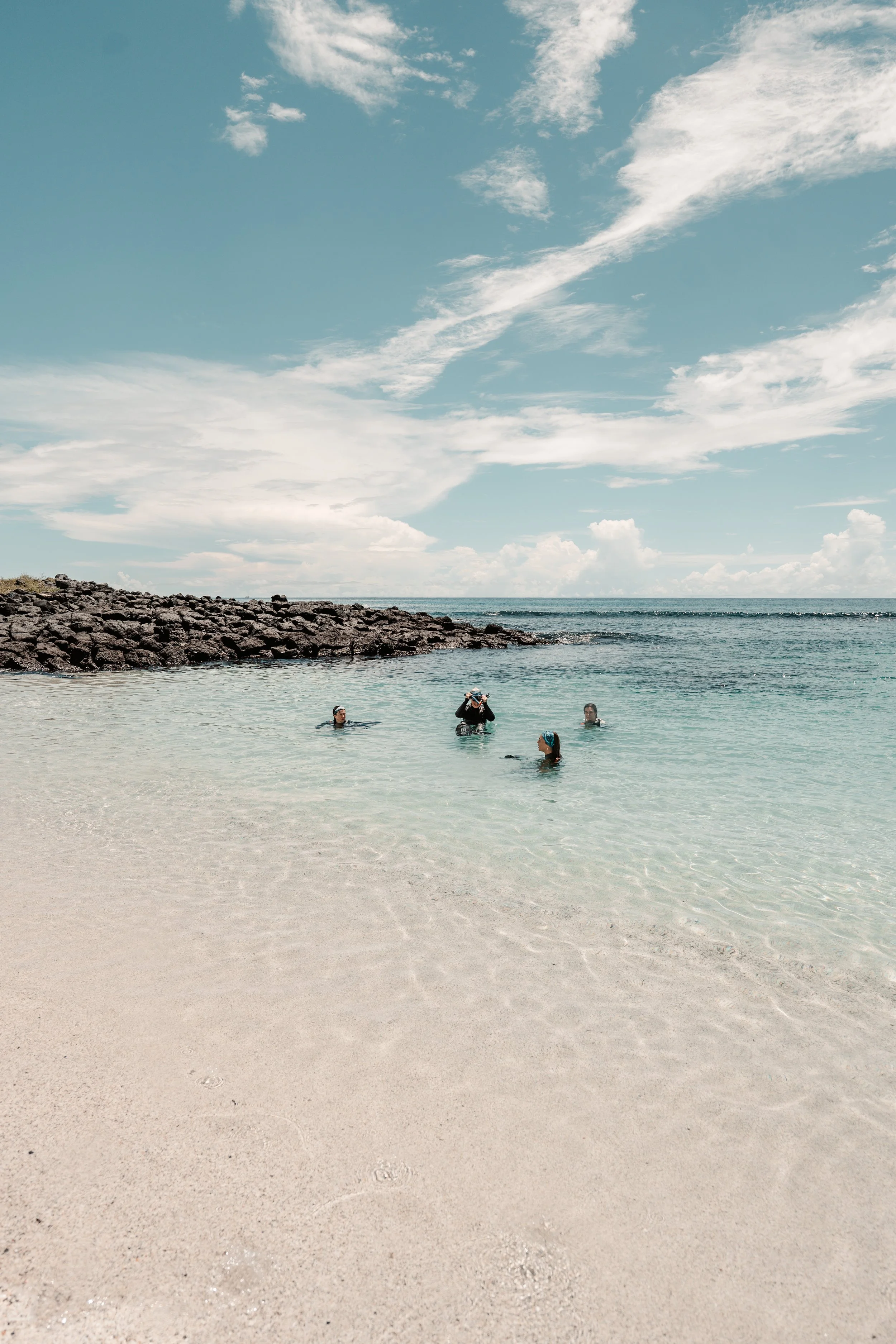 People swimming in the ocean near a rocky breakwater under a partly cloudy sky.