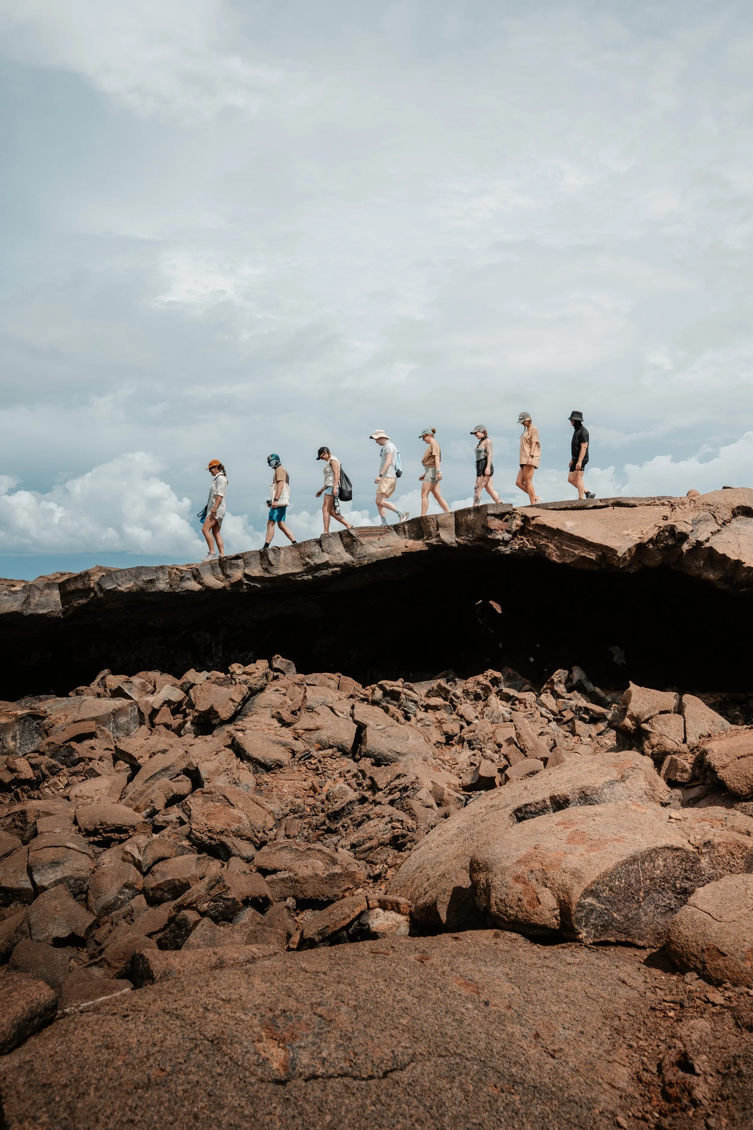 Group of nine hikers walking along a rocky trail under a cloudy sky. Volcanic lava fields at Punta Pucuna, Galápagos.