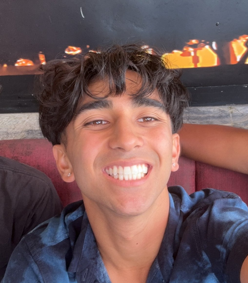 Young man with dark curly hair smiling widely for a selfie, wearing a blue shirt, sitting on a red couch with a blurry black-and-orange background.