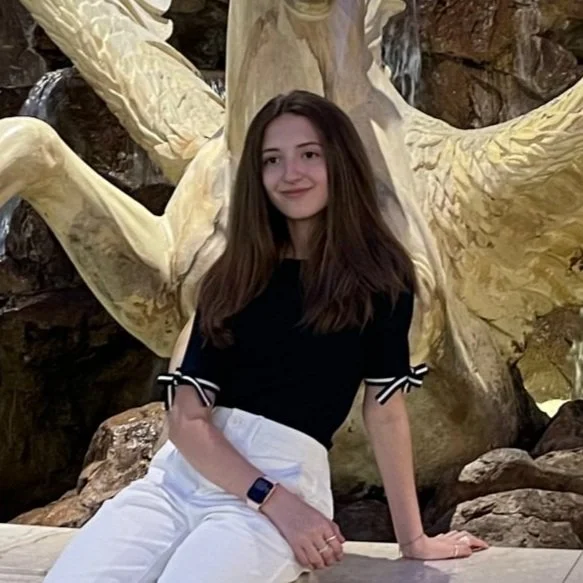 A young woman with long brown hair, wearing a black top with ribbon details on sleeves and white pants, sitting in front of a large decorative sculpture resembling a tree with golden branches.